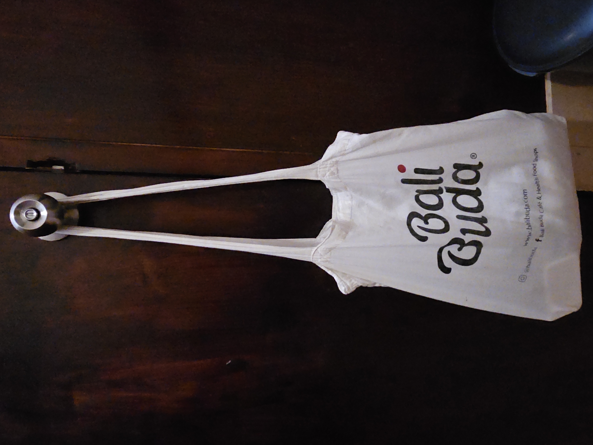 A white tote bag with the words "Bali Buda" printed in black is hanging on a doorknob against a dark wood door. The bag also includes a website and social media handles. The location is likely Denpasar, Indonesia, based on the bag's branding.