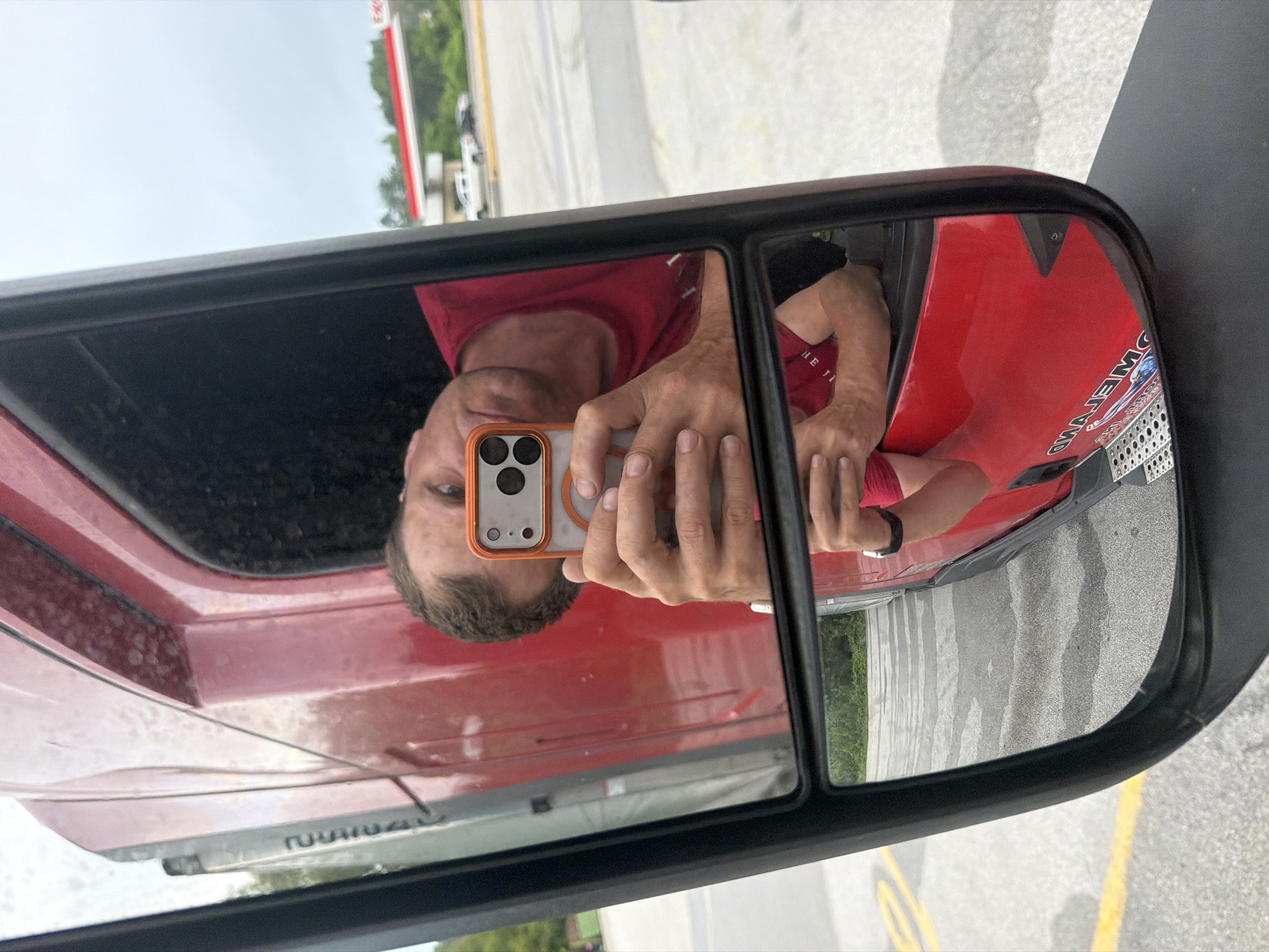 A man in a red t-shirt takes a selfie in a truck's side mirror. The mirror is black and rectangular, with a smaller rectangular mirror below it. The man's face, arms, and chest are visible in the upper mirror, as is his phone. In the lower mirror, the reflection of the truck's red body and the man's torso are visible. The background shows a gas station with a white truck and a red and white sign, trees, and a cloudy sky. The ground is paved. The man appears to be in the city of Wildwood, United States, as indicated by the gas station sign and the general landscape. The overall scene is a casual snapshot taken by the man while he is likely at a truck stop or rest area.
