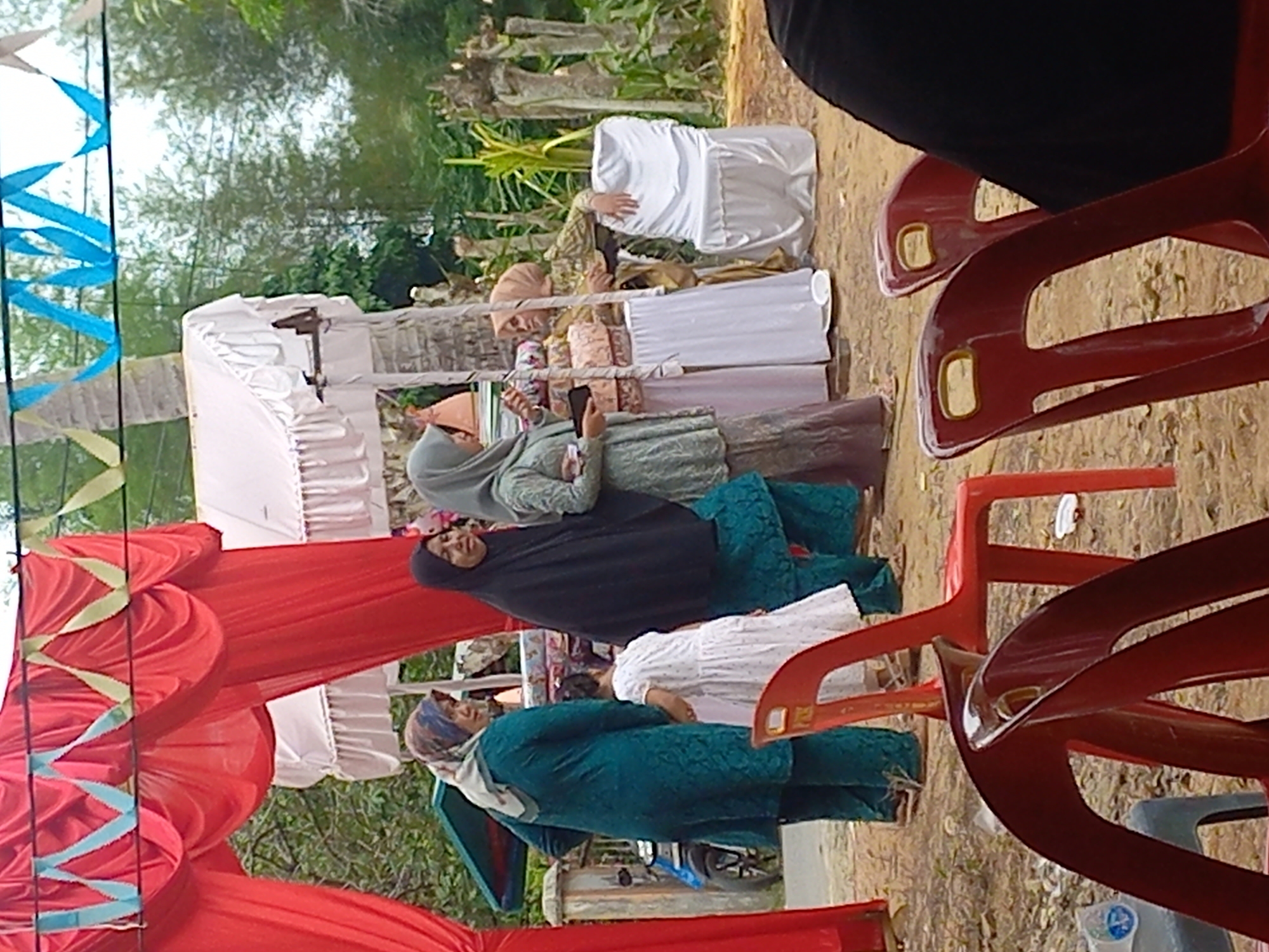In Gampongkeude, Indonesia, a group of people are gathered outdoors under a red fabric canopy adorned with blue and yellow ribbon decorations. Several women, some wearing hijabs, are present. One woman in a teal outfit stands near a child in a white dress. Another woman in a black hijab stands beside her. Further back, a woman in a light green outfit is holding a black object, possibly a phone, and interacting with another woman in a tan hijab who is leaning on a table covered with white fabric and decorative items. To the right, a white-covered chair is visible. In the foreground, red plastic chairs are arranged. The environment appears to be a dirt-covered area with trees in the background. A motorcycle is partially visible to the left.
