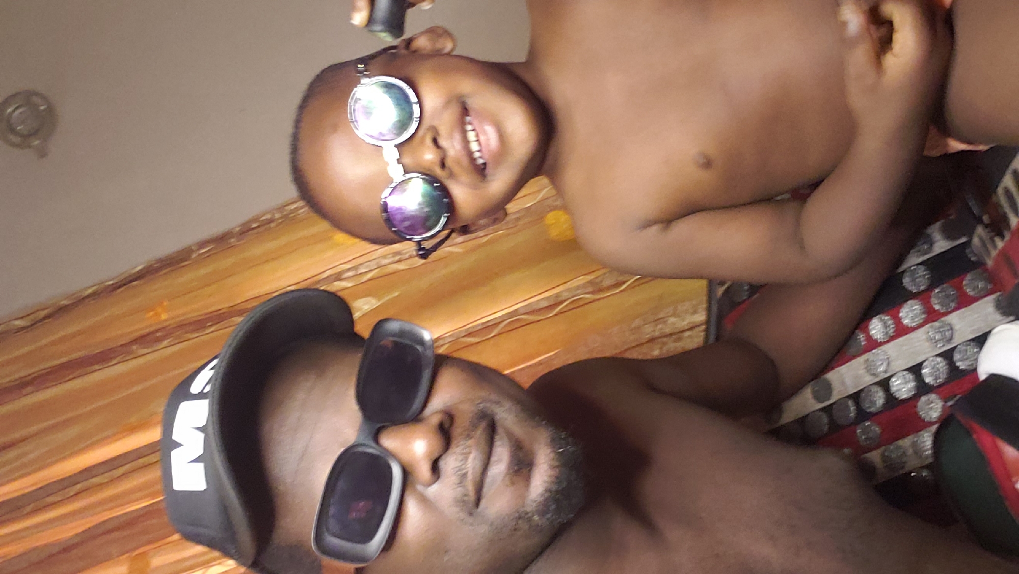 The media file captures an intimate indoor portrait featuring an adult male and a young child, both with dark complexions. The image is oriented sideways, suggesting the subjects are lying down and looking upwards towards the camera.
The adult male, positioned closer to the foreground, is shirtless and has a short beard. He is wearing a black baseball cap with a white letter or symbol (possibly an 'M') visible on the front, and dark square-framed sunglasses that obscure his eyes. His expression is neutral.
Beside him, a young child, also shirtless, smiles broadly, revealing their teeth. The child wears round, reflective sunglasses with a thin silver frame. Their left arm is visible, and they appear to be resting or playing.
The background consists of two main elements: a patterned fabric surface and a wooden-textured surface. The patterned fabric, possibly a bedspread or sofa cover, features horizontal stripes in red, black, and white/silver with circular patterns. The wooden surface, with a reddish-orange hue and visible grain, is likely a floor or piece of furniture. A small, light-colored fixture is visible on the upper wall or ceiling in the background.
The overall scene conveys a candid and joyful moment between the two individuals, likely within a home setting in Sabon Tasha, Nigeria.