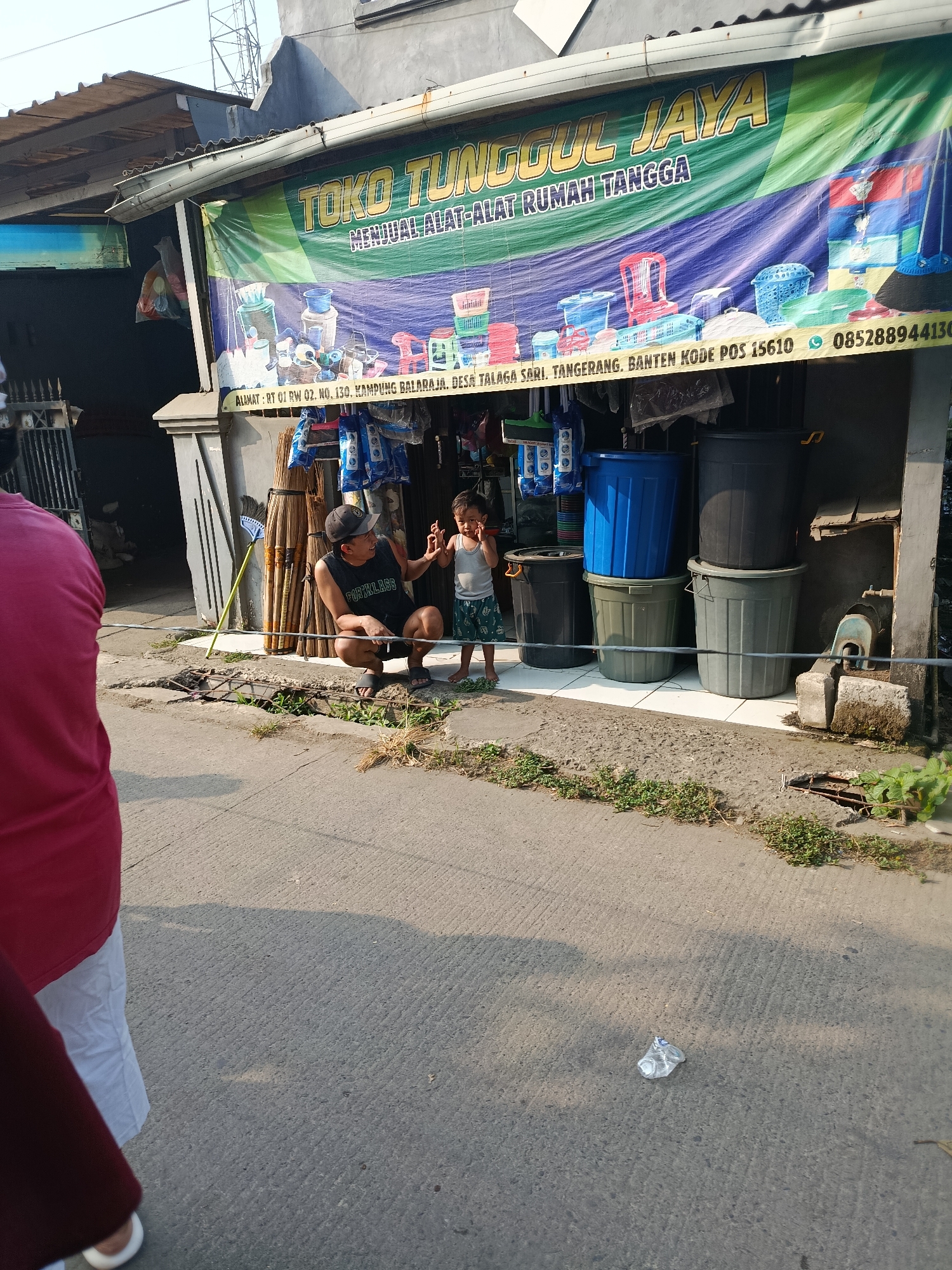 In Balaraja, Indonesia, outside a store called "TOKO TUNGGUL JAYA" which sells household goods, a man wearing a black t-shirt with "FORKLASS" printed on it and a baseball cap squats down. He is interacting with a young child who is wearing a light gray tank top and patterned shorts. The child stands in front of several large plastic bins of various colors, including blue and black. Behind them, the store displays brooms and other items. The scene is set on a sidewalk with cracked pavement and some overgrown weeds. A crumpled plastic bottle lies on the paved ground in the foreground.