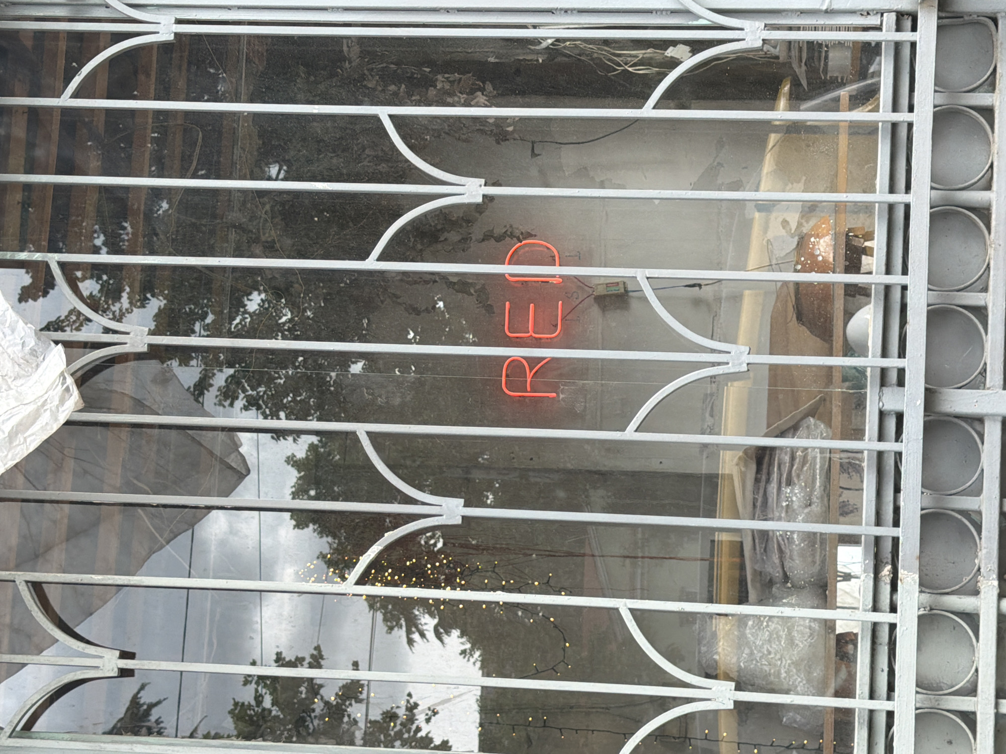 The image shows a barred window with an illuminated "RED" sign inside, viewed from outside. The exterior view through the bars is blurred and reflects trees, sky, and lights. Inside, beyond the sign, are various obscured objects, including what appears to be wrapped items and parts of furniture. The frame of the window is light gray metal, with horizontal bars and decorative curves. The location is likely Tbilisi, Georgia, inferred from the context.