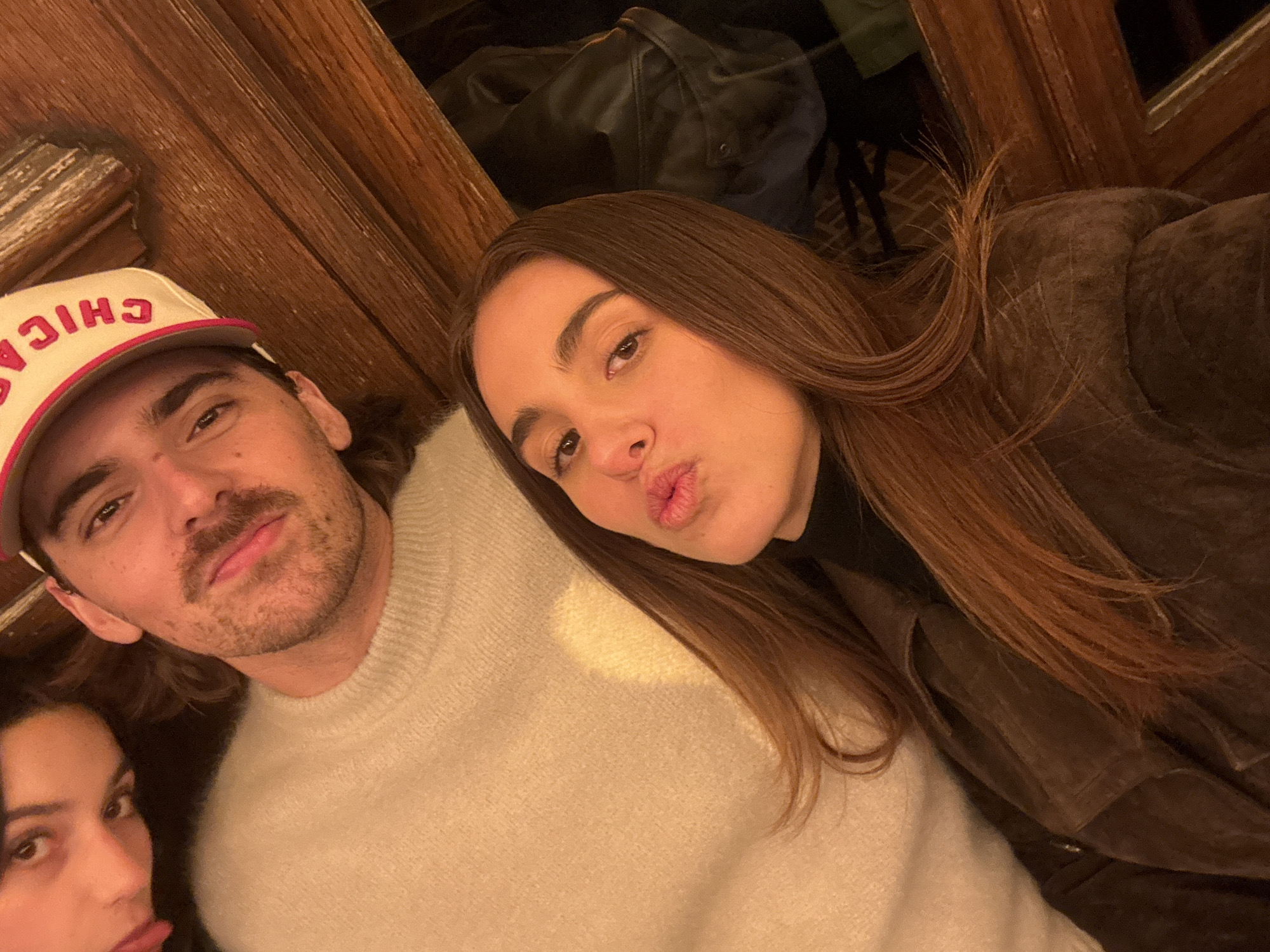 A selfie captures three people. The person on the left is cut off, displaying only a partial view of their face. A man with a mustache wears a "CHICAGO" baseball cap and a cream-colored sweater. A woman with long brown hair is positioned beside him, making a kissing face. The scene appears to be indoors, possibly in a restaurant in Paris, France, suggested by the style of the wood wall paneling. The lighting is warm. In the background, dark-colored objects are visible, potentially jackets or bags hung over the back of a chair.