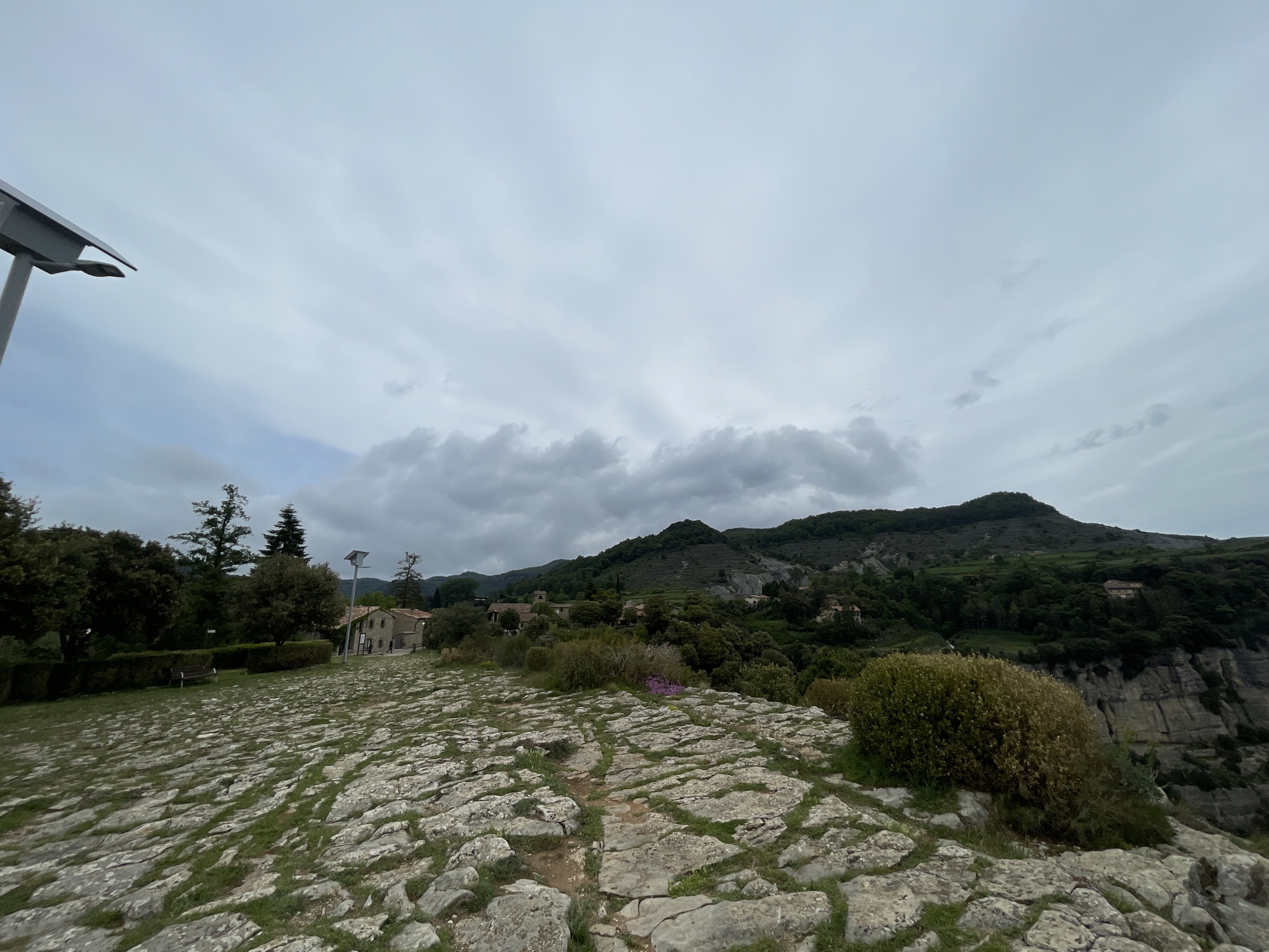 The image depicts an elevated, stone-paved pathway in Tavertet, Spain, under a heavily clouded sky. The path, uneven and cracked, is interspersed with tufts of grass. In the distance, a landscape of rolling hills and rocky outcroppings is visible, punctuated by clusters of trees and a few scattered buildings. Closer to the viewer, shrubs and small patches of purple flowers grow along the edge of the path. A street light is visible in the upper left. The scene suggests a high vantage point, possibly overlooking a valley or gorge, with exposed cliff faces on the right-hand side of the image.
