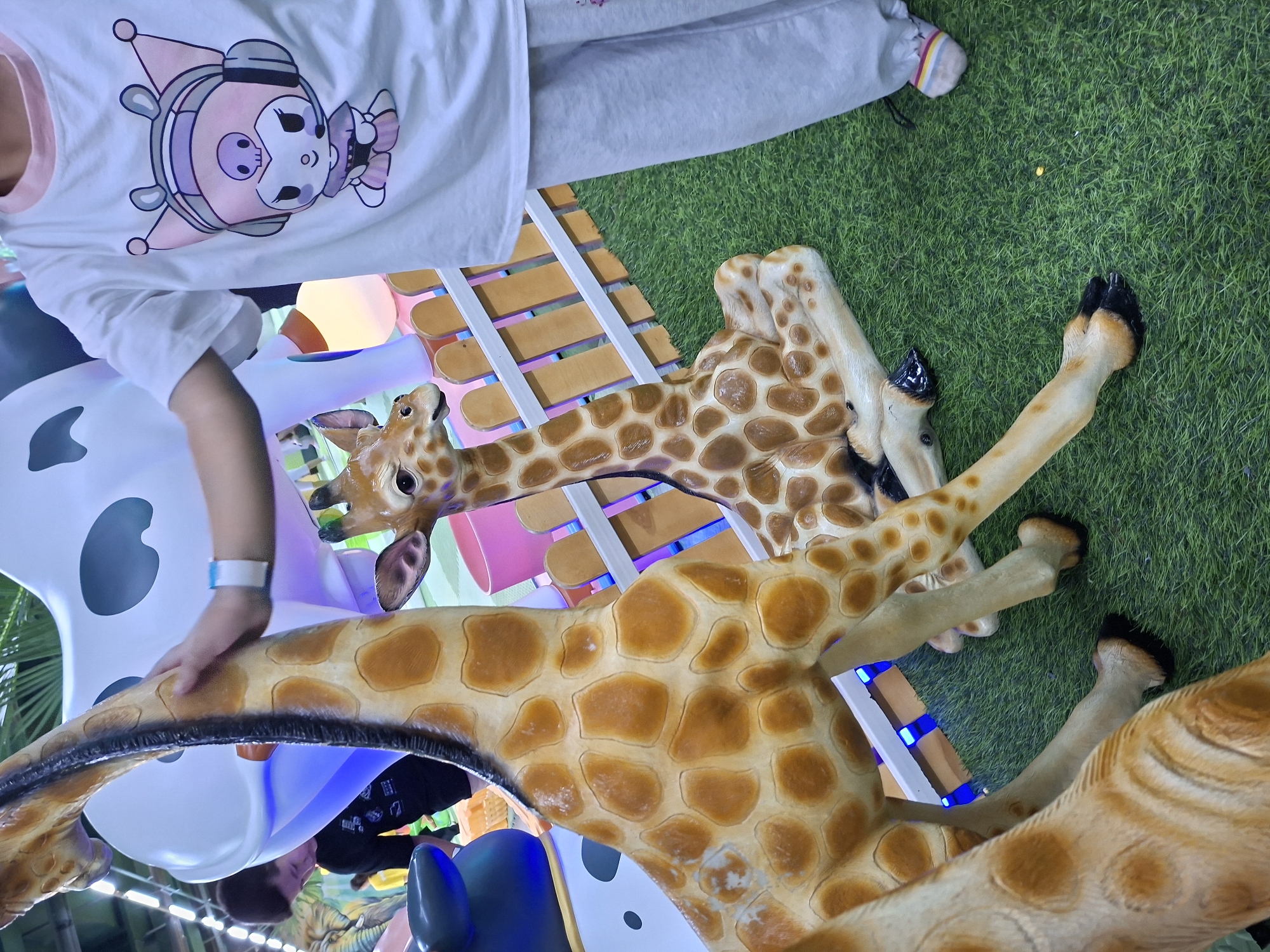 This image depicts a playful indoor setting, possibly an entertainment center or children's play area, in Bishkek City, Kyrgyzstan. The scene features several large, realistic giraffe statues, two of which are prominently in the foreground. The giraffe on the left is partially visible, showing its neck and patterned hide. The giraffe in the center is seated, with its head turned to the right, and its patterned coat is clearly visible. A person, appearing to be a child, is standing to the right of the center giraffe and is reaching out to touch its neck. The child is wearing a white t-shirt with a cartoon character graphic and grey sweatpants. They are also wearing colorful striped socks. In the background, a large white cow statue with black spots is visible, along with other playground elements like a wooden fence and artificial green grass flooring. The lighting in the area is a mix of ambient light and colorful, likely LED, lighting which adds to the playful atmosphere. Another person, a young male, is partially visible in the background on the left, looking down. The overall impression is one of a fun and engaging environment for children.