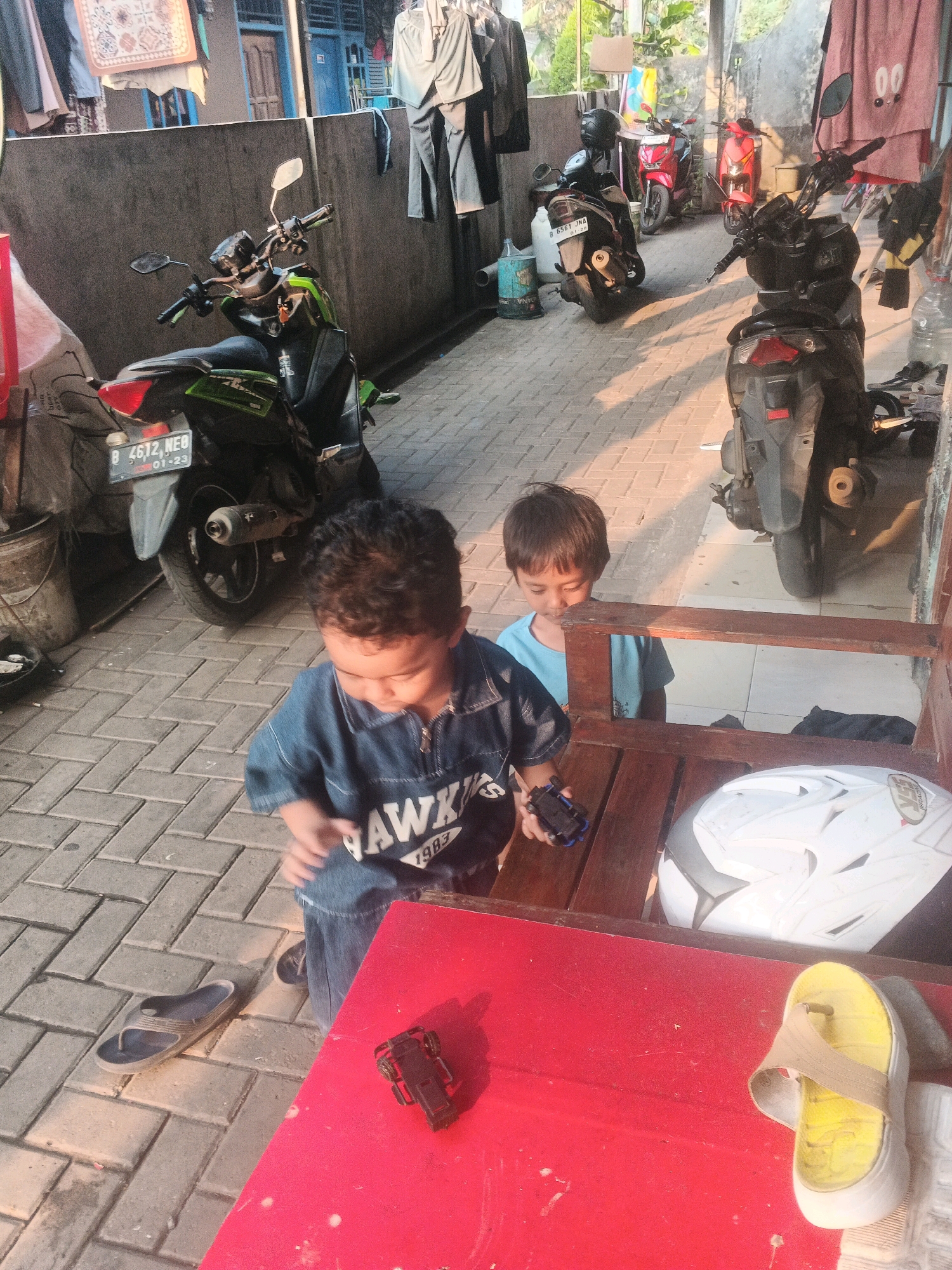 In Kadu, Indonesia, two young boys are in a paved outdoor area with several motorcycles parked behind them. The boy in the foreground, wearing a denim shirt, looks down at a toy car in his hand. He has curly dark hair. The boy behind him, wearing a blue t-shirt, is partially visible. A white motorcycle helmet sits on a red table in front of them, and a yellow sandal is next to it. The area appears to be a residential or communal space, with laundry hanging on a line in the background. The lighting suggests it is late afternoon or early morning.