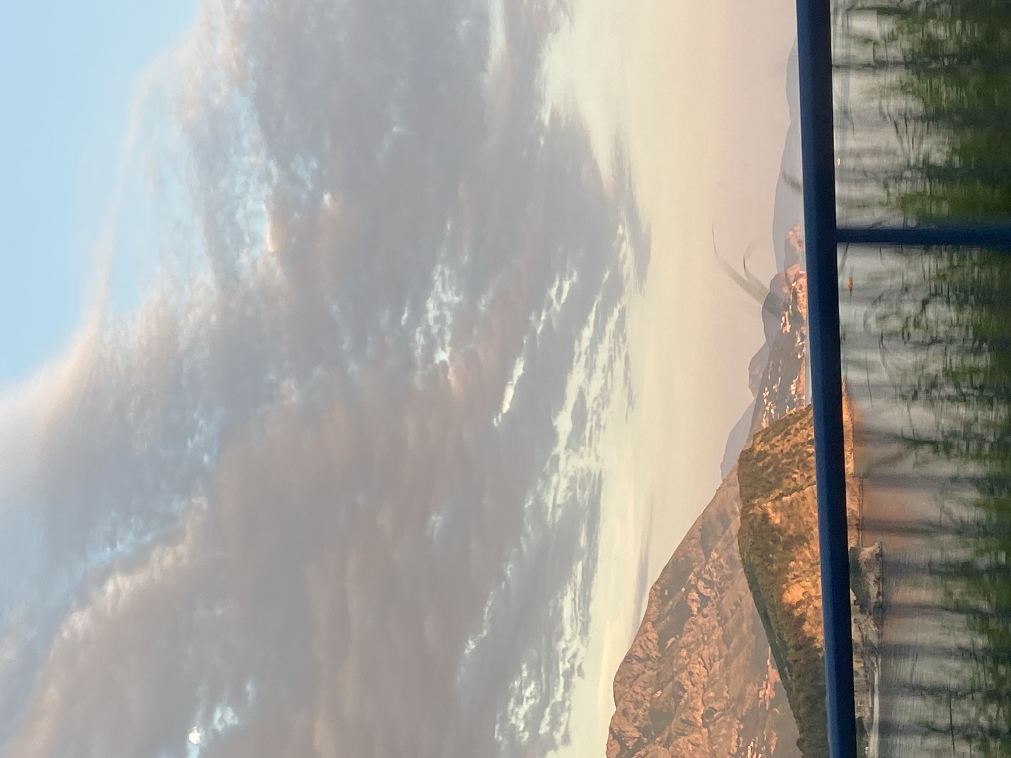 The image, when viewed in landscape orientation, captures a scenic coastal mountain landscape during either sunrise or sunset. The foreground features a dark blue vertical frame on the right, suggesting the photo was taken through a window or from behind a railing, with blurry reflections of green foliage visible behind it.
In the midground and background, majestic mountains dominate the scene, bathed in a warm, golden light that highlights their rugged, rocky terrain and interspersed green vegetation. Distant buildings or settlements can be observed clinging to the mountain slopes, indicating human presence.
The sky is expansive, transitioning from a pale blue at the top to a softer, hazy orange-white hue near the horizon. Scattered clouds, varying in density and texture, are visible across the sky, catching the warm light with shades of white, grey, and subtle orange. A narrow body of water, likely a sea or bay, stretches across the lower-mid section of the frame, reflecting the sky and distant light. The overall impression is one of tranquil natural beauty under the atmospheric glow of twilight. No specific activity or visible text is present. The landscape is consistent with the mountainous coastline of Budva, Montenegro.