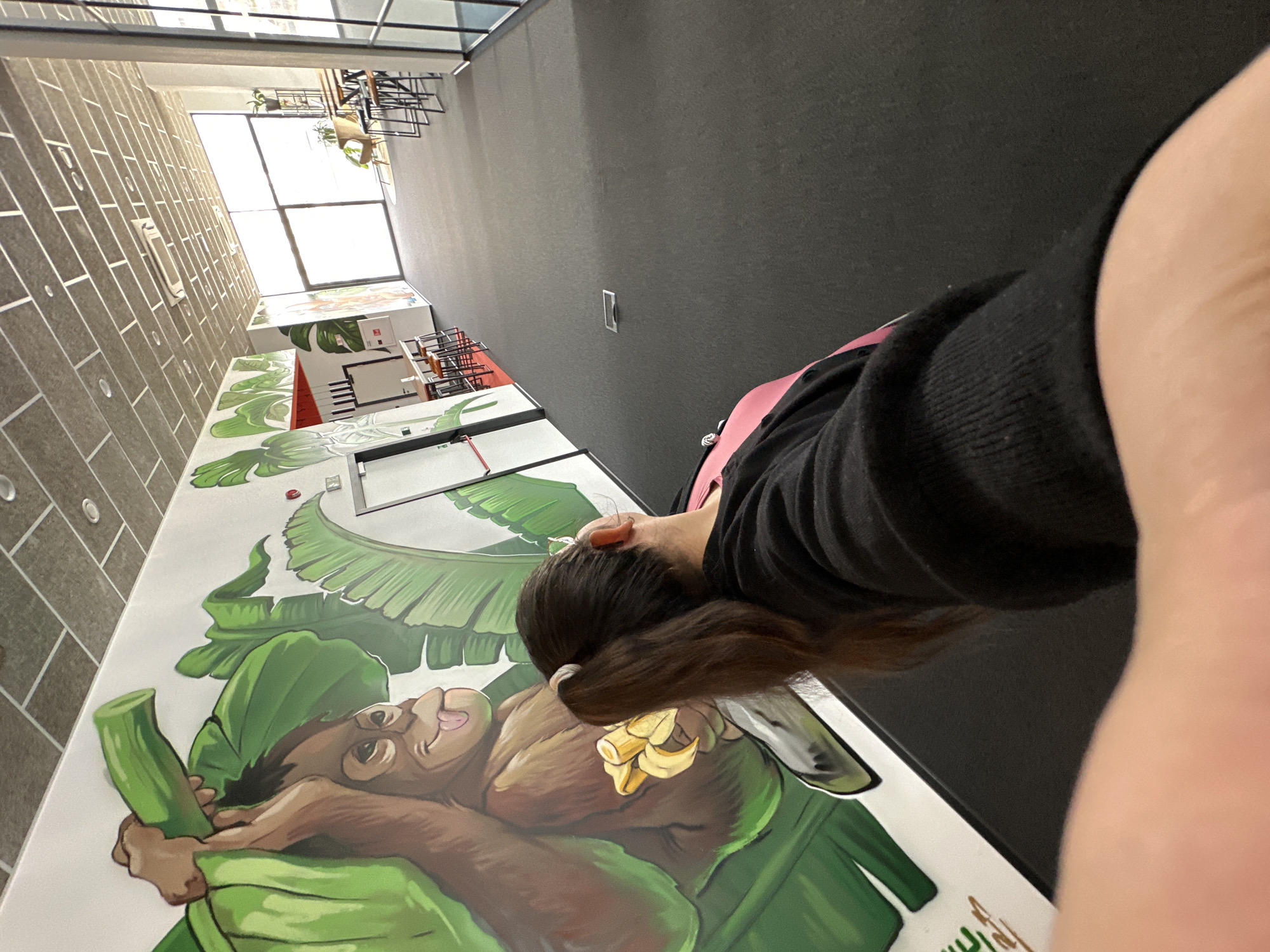 Here's a detailed description of the image:
**Scene:** The image appears to be taken inside a modern building or office space, potentially in Varna, Bulgaria.
**Environment:**
* The building has a high ceiling and ample natural light coming from large windows.
* One wall is covered in light-colored brick, while another is gray.
* A vibrant mural depicting an orangutan holding bananas and surrounded by large green leaves is visible on a surface, possibly a table or a wall.
* The interior design is contemporary.
**People:**
* A person is partially visible, mostly showing their hair, part of their face, and the back of their black sweater with a pink underlayer.
**Objects:**
* A mural with an orangutan eating bananas.
* Bananas.
* Leaves.
**Actions and Interactions:**
* The person may be looking at or interacting with the mural.