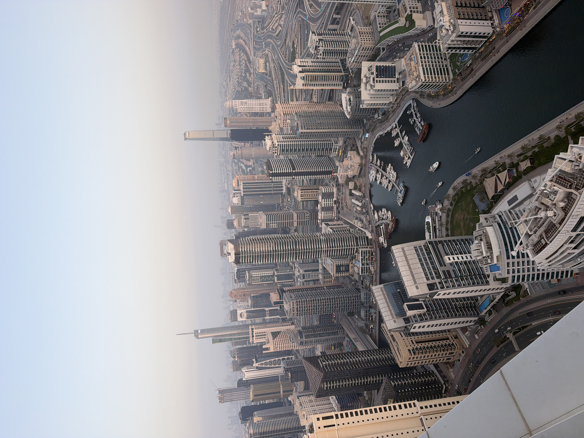An aerial view of Dubai Marina, United Arab Emirates, displays numerous high-rise buildings densely packed together. A waterway flows through the center of the urban landscape, featuring several boats and yachts docked along its edges. Highways with vehicular traffic weave among the buildings, and in the distance, the city fades into a hazy sky. The photograph is taken from a high vantage point, possibly from the top of another skyscraper.