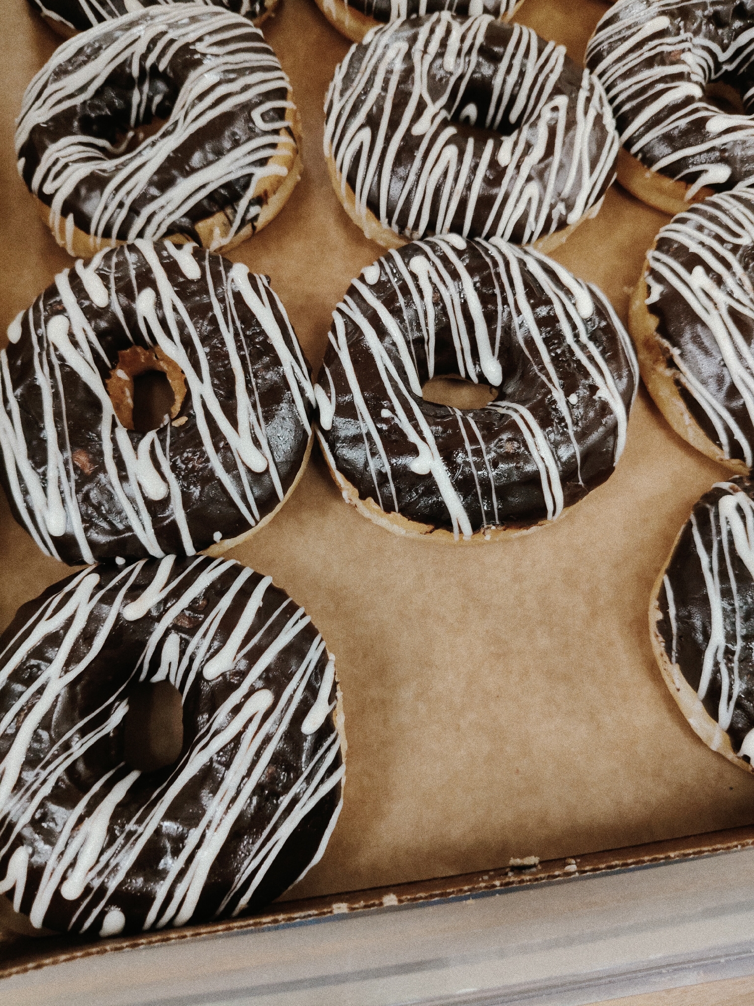The media file shows a close-up, overhead view of several chocolate-frosted donuts with white icing drizzles arranged in a box. The donuts appear to be freshly made, with a glossy chocolate glaze and a pattern of thin white stripes. The box is lined with brown paper, giving it a rustic feel. The lighting is soft and diffused, suggesting an indoor setting. The donuts are the sole focus of the image, with no people or other objects visible. The overall impression is one of a tempting treat, evoking feelings of indulgence and satisfaction. The location is Kemerovo, Russia, though no specific landmarks or cues indicate this within the image itself. There is no visible text in the media file.
