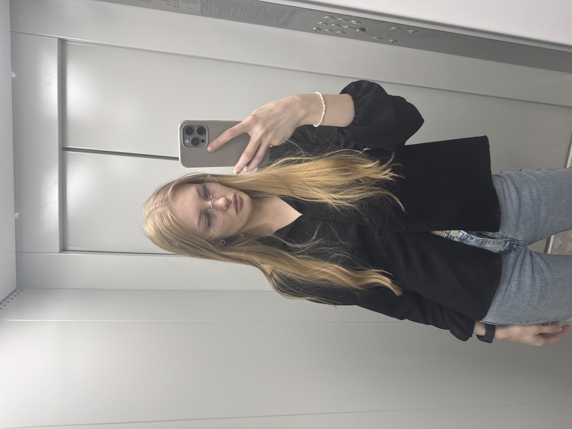 The media file is a selfie taken by a young woman in an elevator. She has blonde hair, glasses, and is wearing a black blouse and light blue jeans. She is holding a smartphone in her right hand to take the photo. On her left wrist, she is wearing a pearl bracelet. The elevator has gray walls and a metal panel with buttons. The time of day and weather are not discernible from the image. The location is identified as Perm, Russia, which could be inferred from metadata not present in the visual information. No visible text is present in the image. The woman's expression appears neutral.