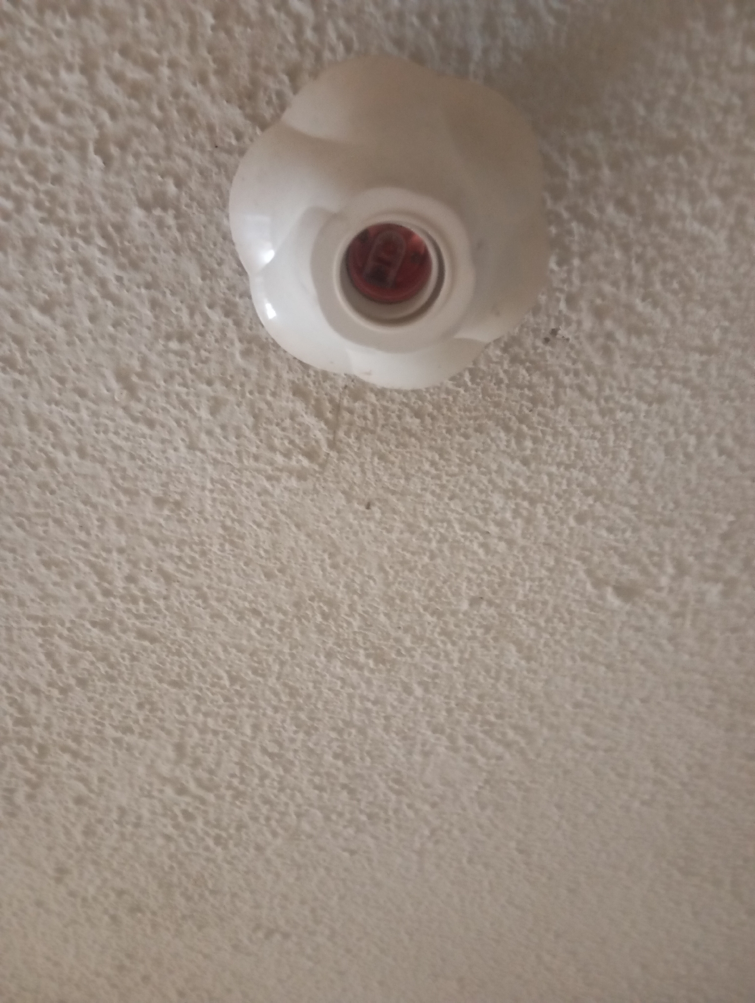 A white flower-shaped light fixture is mounted on a textured white ceiling. The fixture has a red interior where a bulb would be inserted. The ceiling's rough texture is visible throughout the image, suggesting it may be a popcorn or stucco finish common in older buildings. The lighting in the image is overhead, casting a soft light that illuminates the ceiling and the fixture. There are no people or other objects visible in the scene. This scene is generic and could be found in any building with this type of ceiling and light fixture, making it impossible to pinpoint a specific location within Jos, Nigeria, or any other city.