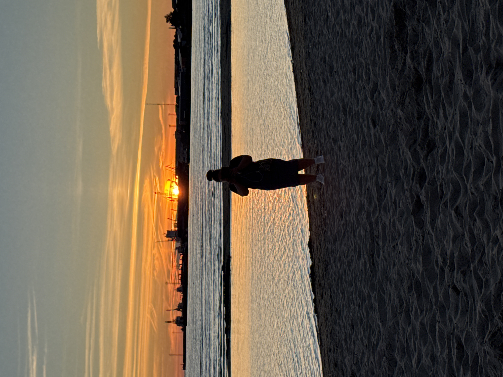 A silhouette of a person stands on a sandy beach with the Baltic Sea in the background. The sun is setting, casting an orange and yellow glow across the sky, with the silhouette of industrial cranes and buildings visible on the horizon, suggesting a port area, likely in Tallinn, Estonia. The water reflects the sunset colors. The person is facing the water and appears to be holding a camera or phone, possibly taking a photo. The sand is dark and textured, with ripples indicating the tide.