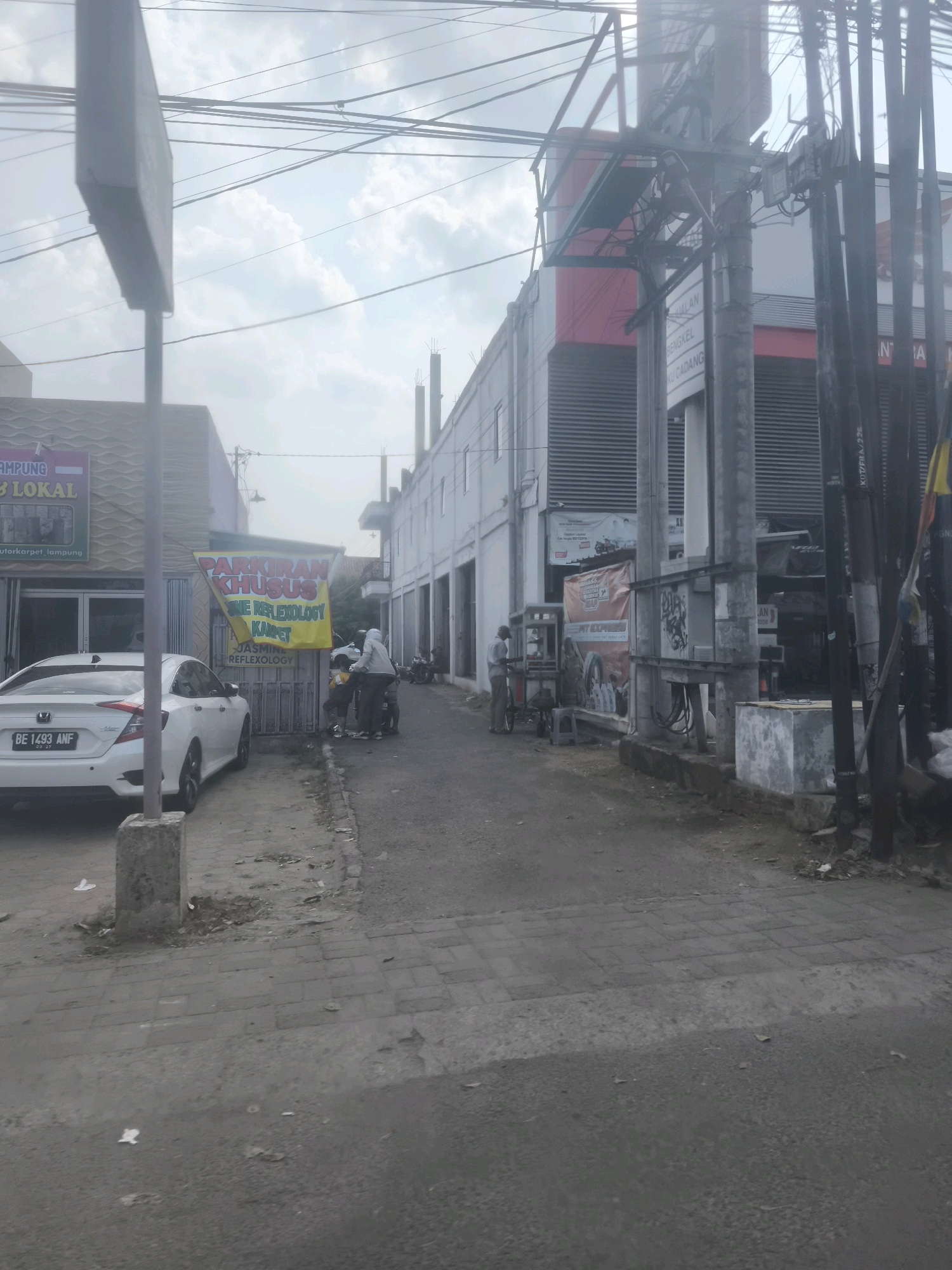 The image captures a street scene in Bandar Lampung, Indonesia. In the foreground, a white Honda Civic sedan with license plate BE 1493 ANF is parked on the left side of the road. A grey concrete post is positioned near the car. The street is paved with grey asphalt and bordered by a sidewalk made of interlocking grey pavers.
Further down the street, a yellow banner with red lettering reads "PARKIRAN KHUSUS" (Special Parking) and "JASMINE REFLEXOLOGY KARPET". Several people are gathered near a motorbike, some appearing to be talking or standing. A few other motorbikes are parked in the vicinity.
On the right side of the street, a white building with grey horizontal siding dominates the background, with some red accents on its upper facade. Signs with Indonesian text are visible on this building, including one that appears to read "BUKA" (Open). Utility poles with numerous wires crisscross the sky, typical of urban electrical infrastructure. The sky is overcast with clouds. To the far right, a section of a red and yellow striped flag is visible. The overall impression is that of a commercial street with shops and services.
