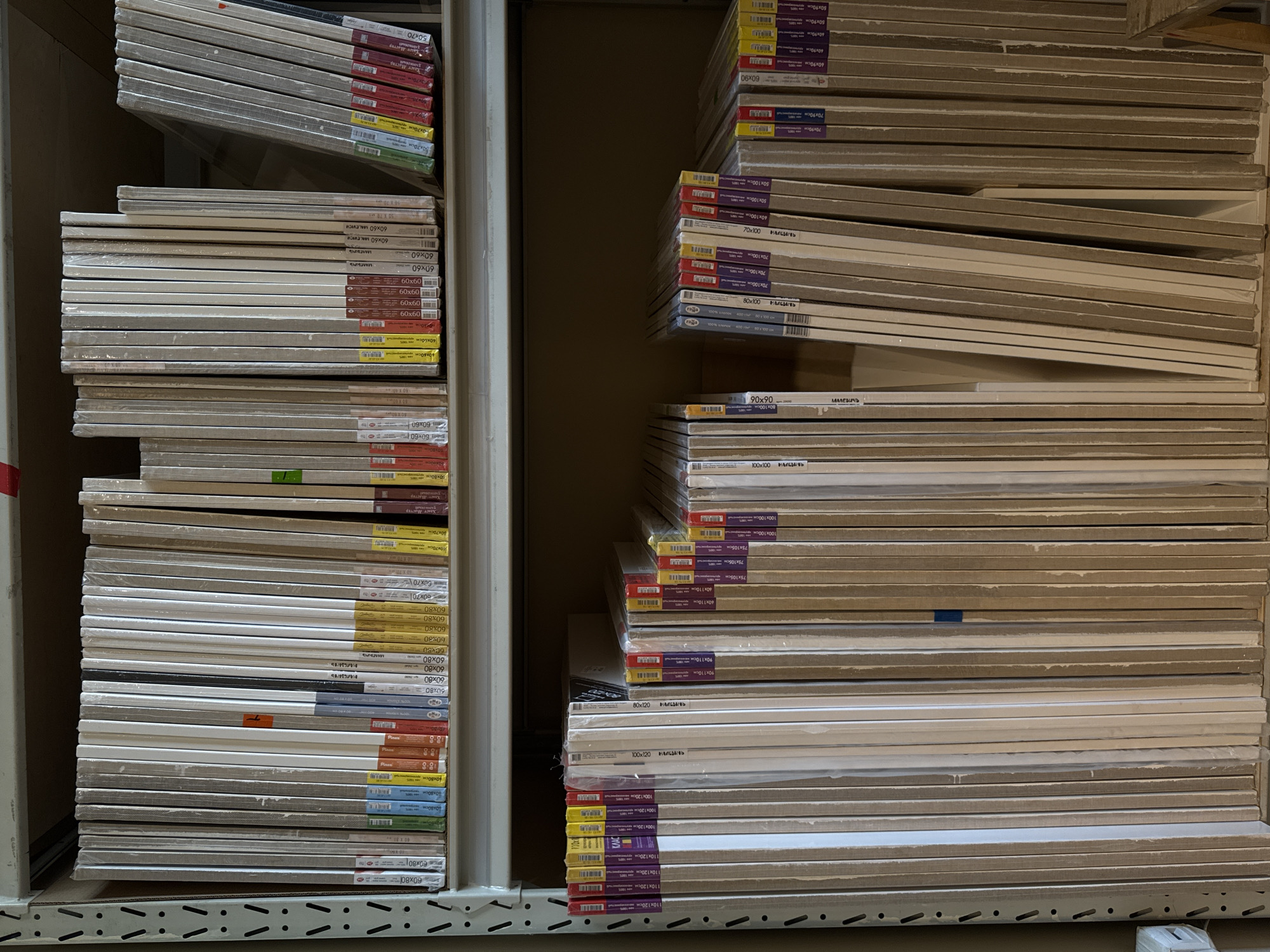 This is a photograph of a retail display of canvases for painting. The canvases are stacked on shelves, some are standing upright, while others are leaning. They are mostly packaged in clear plastic wrap, with colorful labels indicating their size and brand.
The subjects are the canvases themselves, which are the primary focus of the image. They are of various sizes, ranging from smaller dimensions like 50x70cm and 60x60cm to larger ones such as 100x120cm and 110x120cm. The brand "МАЛЕВИЧЬ" (Malevich) and "LALEVICH" are clearly visible on several of the packages. Other text visible includes dimensions in centimeters (e.g., "60x80cm," "70x90cm," "90x110cm") and some Russian words that appear to be related to the art supplies, such as "Хаст Мастер" (Art Master) and "рисий" (possibly Russian). There are also labels with "ONO" and "raja."
The scene is indoors, likely in a store or a warehouse, given the shelving units and the organized arrangement of the products. The lighting is artificial, creating a slightly dim and shadowed atmosphere. There are no people visible in the image. The activity is essentially the display and storage of art supplies. The setting is a commercial space dedicated to art materials. The overall impression is one of a well-stocked art supply store, with a focus on the variety and presentation of the canvases. The colors of the labels add some visual interest to the otherwise neutral tones of the canvases and shelving.