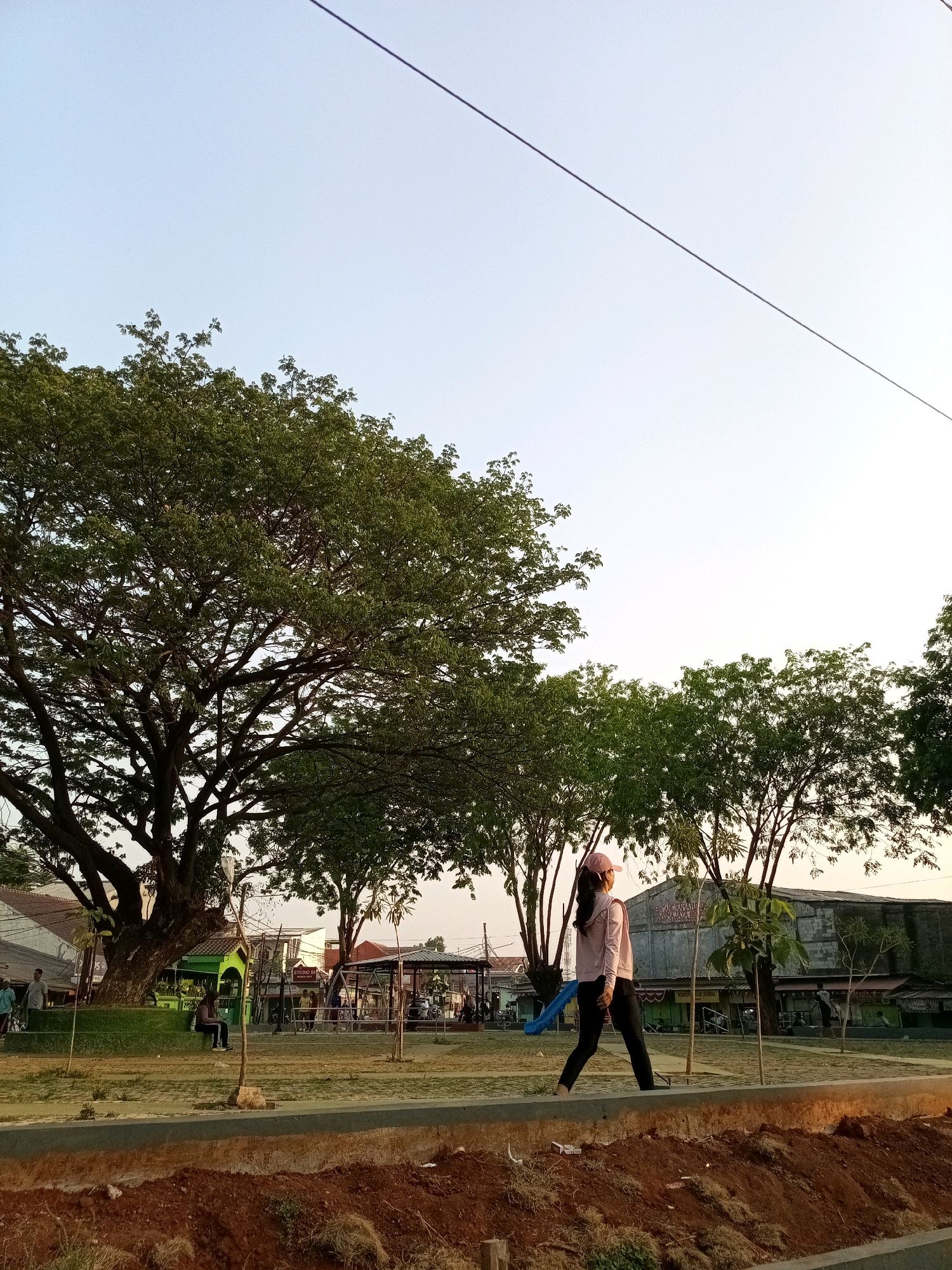 A young woman wearing a pink baseball cap, a pink long-sleeved shirt, and black pants walks across a paved area in what appears to be a park in Bekasi, Indonesia. She is carrying a blue object in her right hand. The park features large trees, including a prominent, mature tree on the left side of the image. In the background, there are buildings, a playground with a blue slide, and a few other people. The sky is clear and bright, suggesting it is daytime, possibly late afternoon. The foreground shows an area of exposed soil with some dry grass. A wire runs diagonally across the upper portion of the image.