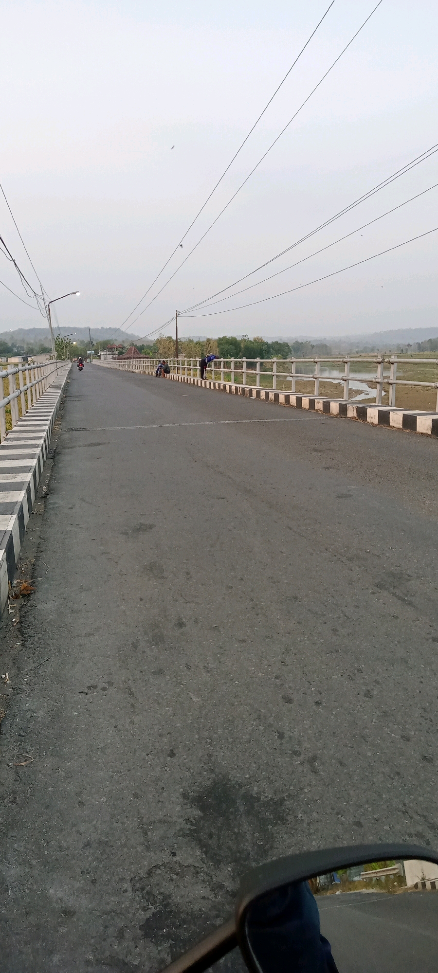 The image depicts a paved road on a bridge in Kemusu, Indonesia. The road is lined with a white and black striped barrier. Several people are visible on the bridge, some standing near the railing and others sitting. In the distance, there is a body of water and lush green vegetation. The sky is overcast. The camera perspective suggests the photo was taken from a moving vehicle, possibly a motorcycle, as a rearview mirror is visible in the foreground.