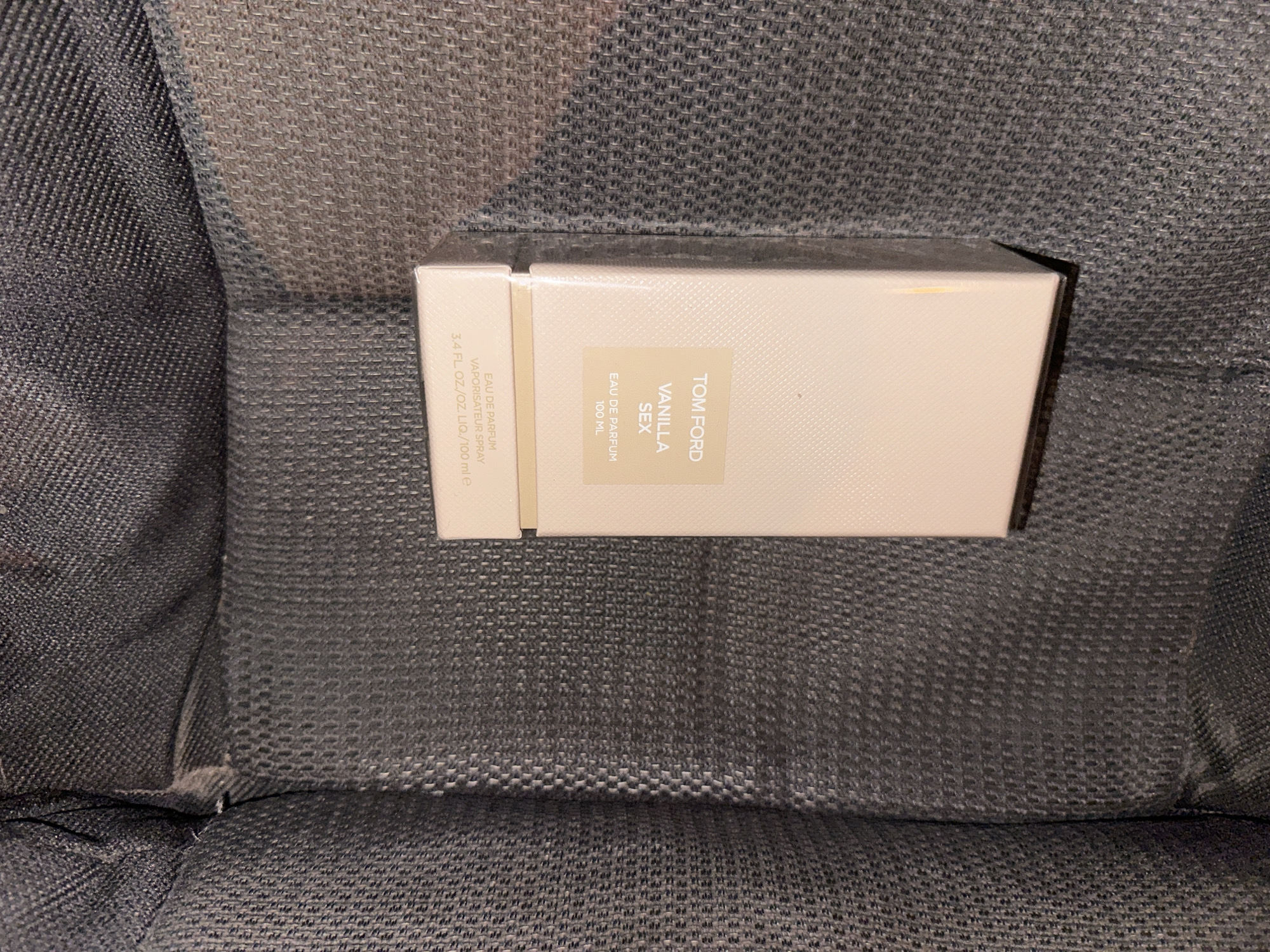 A Tom Ford Vanilla Sex Eau de Parfum fragrance box is displayed on a textured, dark gray fabric surface, possibly upholstery. The box is light pink with black text and features the brand name "TOM FORD" in black, followed by "VANILLA SEX" in a larger, bolder font. Below this, it reads "EAU DE PARFUM" and "100 ML". A second line of text at the bottom of the box states "EAU DE PARFUM VAPORISATEUR SPRAY 3.4 FL. OZ./.OZ. LIQ./100 ml e". The image is taken from a slightly elevated angle, with the box placed centrally. There is no indication of the city of Sliven, Bulgaria, or any other specific location.
