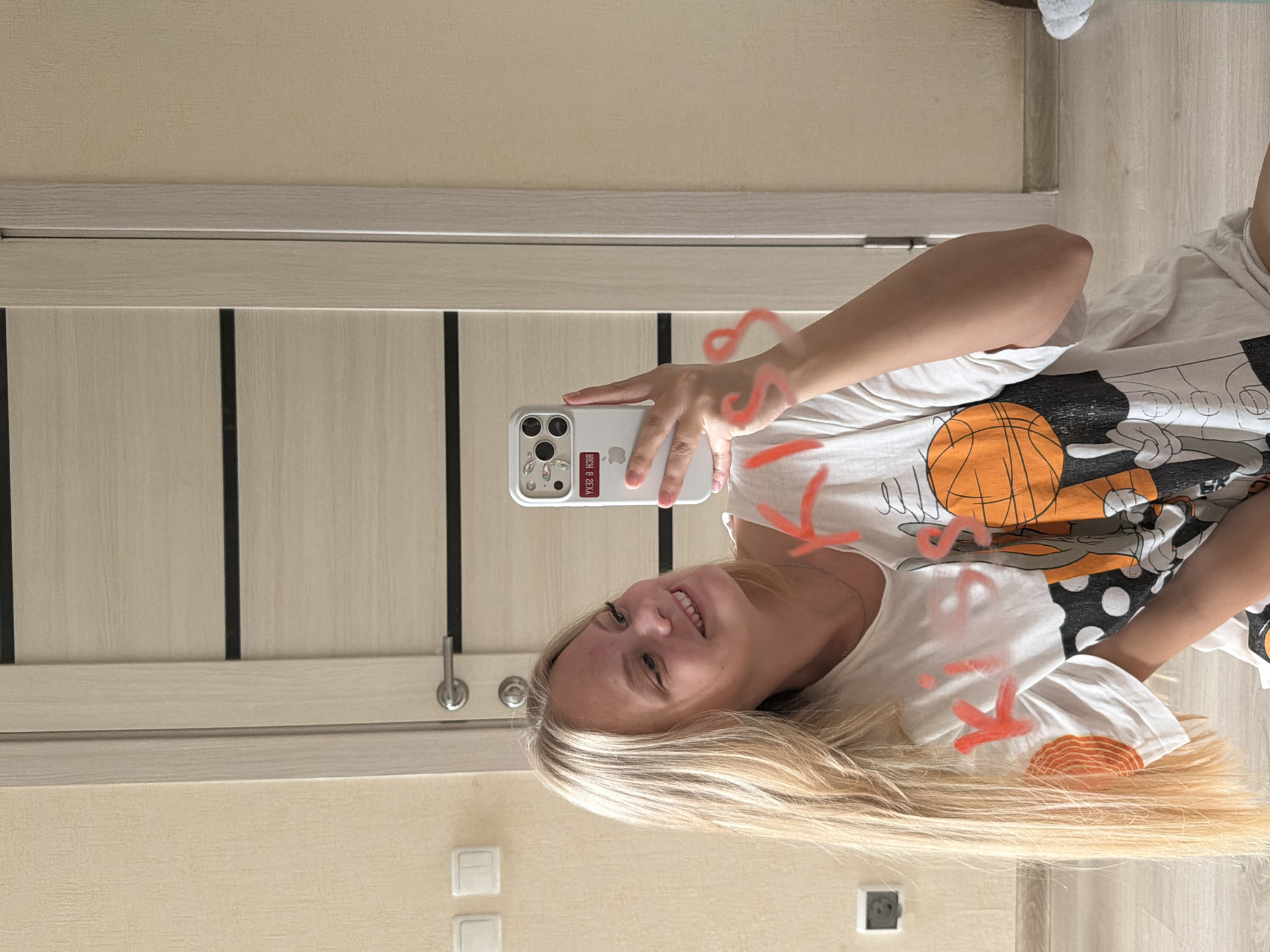 The media file is a vertical, slightly blurry, selfie taken by a young Caucasian woman with blonde hair. She is smiling and looking directly at the camera, holding a white iPhone in her right hand. The phone case has a decorative element resembling rhinestones. She is wearing a white t-shirt with a graphic print featuring cartoon characters and a basketball. The background appears to be an indoor setting, possibly a room in a residential building, with a light-colored wall and a wooden door. There is a horizontal silver trim line across the door. Two electrical outlets are visible on the wall. The lighting suggests it is daytime, but the specific time of day is not discernible. The overall mood is casual and cheerful. Some red scribbles, possibly a filter effect, are overlaid on the image, particularly on the woman's clothing and hair. Based on the provided location context, this scene is likely in Izhevsk, Russia. No other significant text is visible in the image besides what is on the phone screen, which is partially obscured and unreadable.
