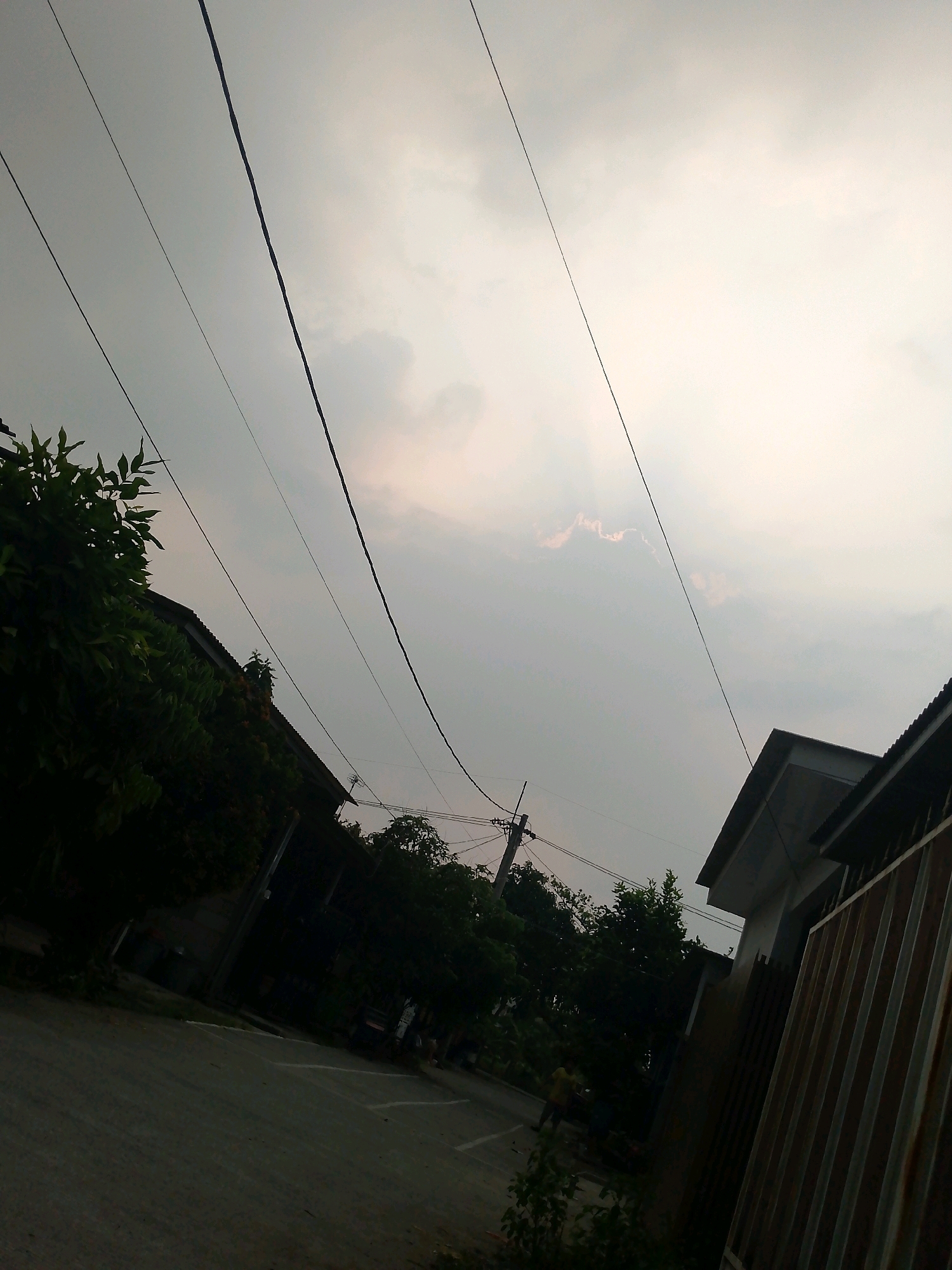 This image depicts a street scene in Klapanunggal, Indonesia. The sky is overcast with diffused light, suggesting either dawn, dusk, or a heavily clouded day. Several power lines crisscross the frame diagonally. On the left side, the dark silhouette of a building covered in dense green foliage is visible. The street itself is paved and appears to be a residential or light commercial area. To the right, another building with a peaked roof and a corrugated metal fence is present. The overall impression is of a quiet, possibly residential area under a subdued sky.