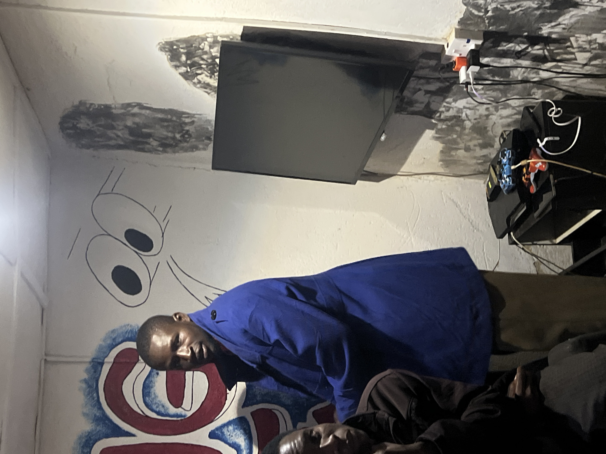 In a dimly lit room with white, textured walls, possibly in Durumi, Nigeria, two individuals are present. A man stands prominently in the center, wearing a bright blue trench coat over khaki pants. He faces partially towards the camera, with an open mouth as if speaking. To his left, another person, largely obscured by shadow, is seated and looking towards the viewer. The wall behind them is adorned with graffiti; to the left, a red and blue design is visible, and to the right, a cartoonish face with large, outlined eyes is drawn. A black flat-screen television is mounted on the wall to the right of the frame, with various cables and a power strip connected below it. Adjacent to the power strip, a black console with two video game controllers, one blue and one red, sits on a black surface. The overall impression is of a casual gathering or gaming session in a decorated, perhaps personalized, living space.