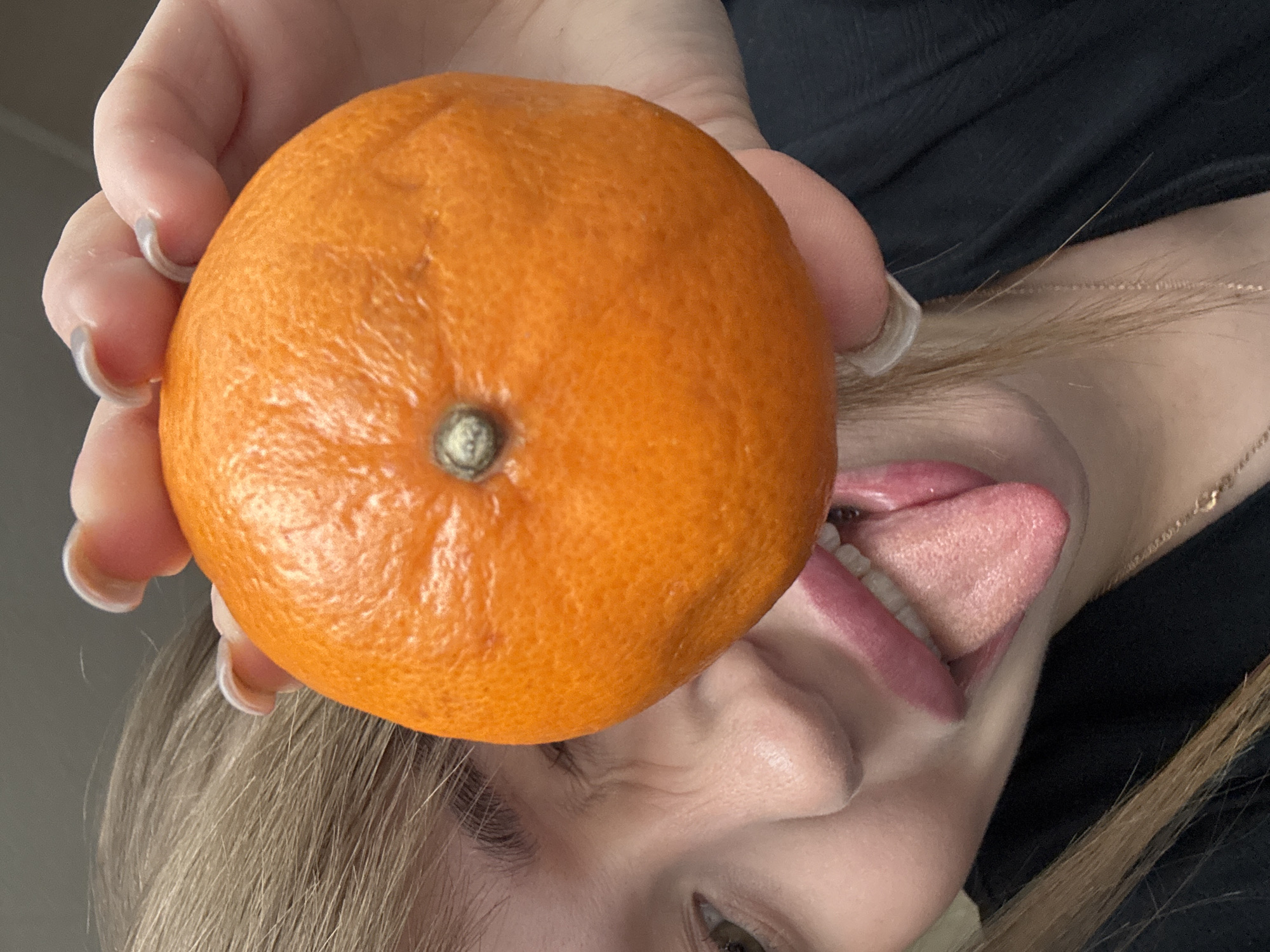 A close-up image shows a person holding an orange in their left hand, with the orange obscuring part of their face. The person is sticking their tongue out towards the orange. The person has blonde hair and light skin, wearing a black shirt and a gold chain around their neck. The fingernails on the left hand are long and white-tipped. The background is a light grey. There is no indication of the location being in Chaikovsky, Russia.
