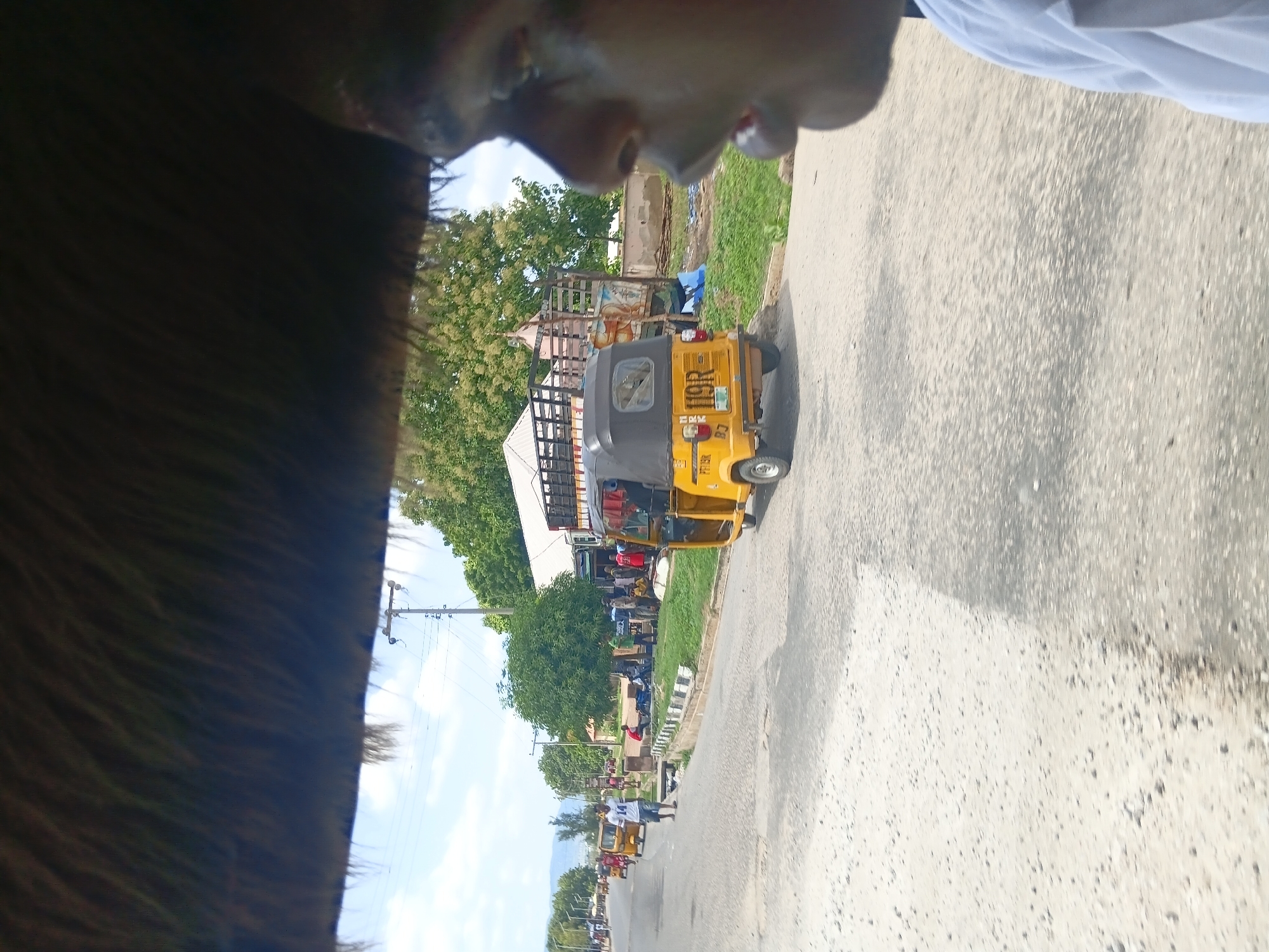 The image depicts a street scene in Jalingo, Nigeria, likely near a market or transportation hub. In the foreground, a portion of a person's face is visible on the right side, with their mouth open. The perspective suggests they are looking to the left. In the middle ground, a yellow auto-rickshaw, or "Keke," is parked on the right side of the road, its license plate reading "119R." Behind the Keke, a large, flatbed truck is parked, appearing to be loaded. Several people are gathered on the left side of the road, some sitting and others standing, near what appear to be market stalls or temporary shelters. Further down the road, several more Keke vehicles are visible, and a few individuals are walking. The environment is characterized by a paved road with visible patches and debris, bordering a grassy area. Trees are abundant in the background, and a utility pole with wires is present. The sky is cloudy. The overall impression is one of a busy, daytime commercial or transportation area.