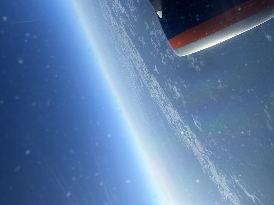 The image presents an aerial view captured from an aircraft, looking downward and towards the distant horizon over a vast landmass. This scene is situated over Russia, with the provided context placing the location above Berezovaya Roshcha. The upper two-thirds of the frame show a clear, deep blue sky, which gradually lightens to a pale blue-white at the horizon line. Scattered across the sky and upper landscape are numerous small, out-of-focus circular white specks, likely particles on the aircraft window. Below the horizon, the terrain consists of extensive dark-colored areas, presumably forests, interspersed with irregular and geometrically shaped lighter patches consistent with snow-covered ground or agricultural clearings. In the lower right corner, a section of an aircraft engine nacelle is visible. It is predominantly dark blue or black, featuring a prominent horizontal orange-red stripe and a metallic silver leading edge. A small white projection, possibly a sensor or probe, extends from the upper surface of the nacelle. No people, specific actions, or direct interactions are visible within the frame.