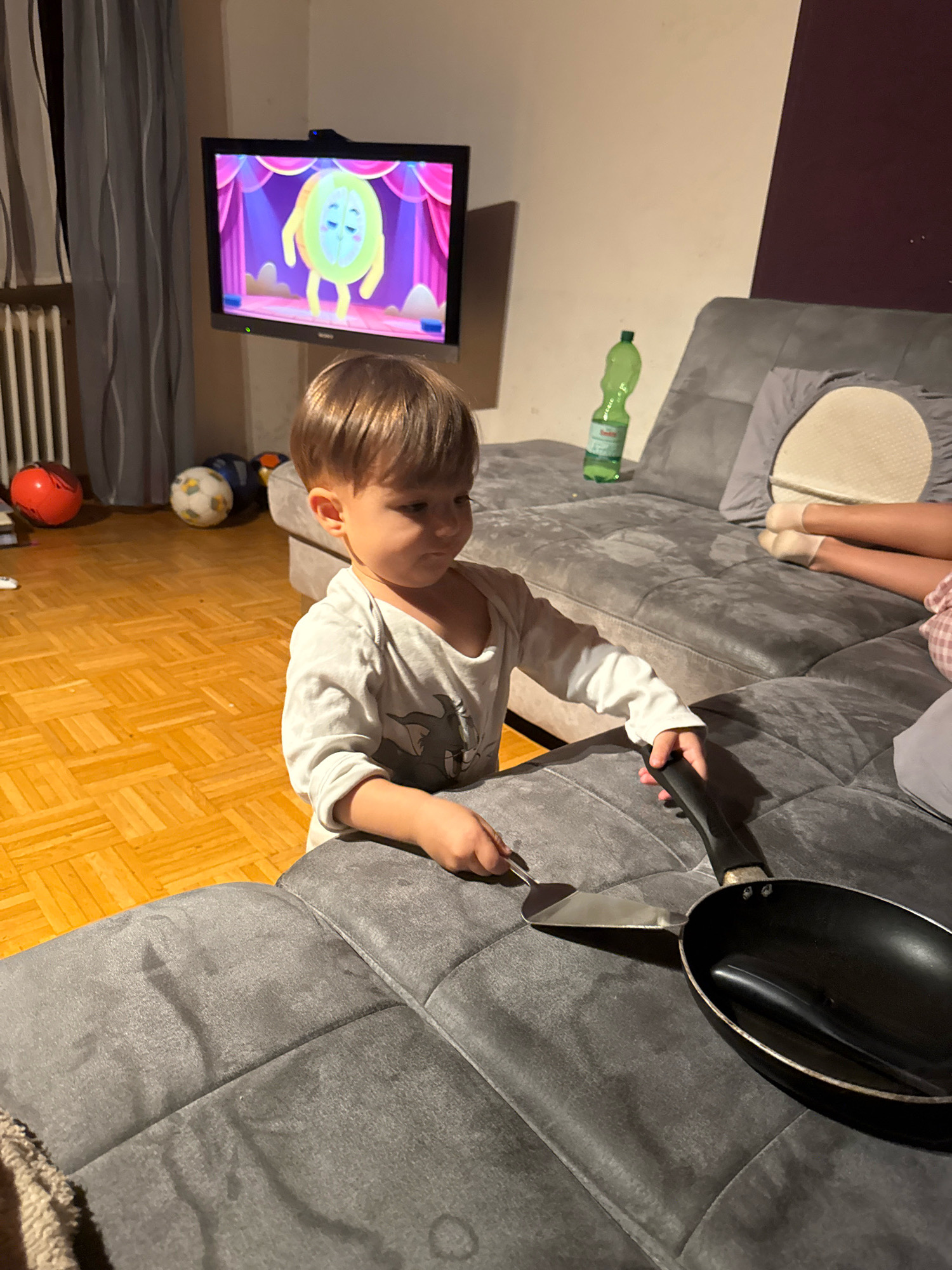 In Überherrn, Germany, a young boy is engrossed in playing with a toy frying pan and spatula on a grey sectional sofa in a living room. The boy, wearing a light-colored long-sleeved shirt, is focused on his pretend cooking activity. Adjacent to the sofa, a television displays a cartoon character. Several soccer balls are scattered on the wooden floor in the background, suggesting a playful environment. A green plastic bottle rests on the sofa, and two pairs of legs, likely belonging to other individuals, are visible further down the couch. The scene is lit by ambient room lighting, creating a relaxed, domestic atmosphere.