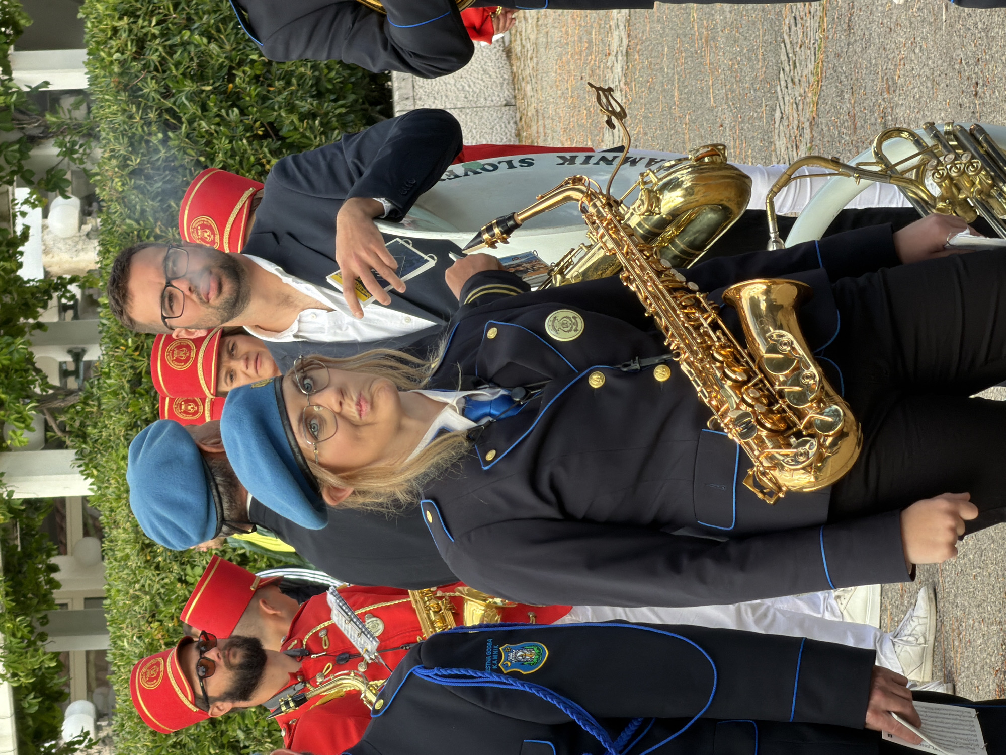 A group of people is standing outside, some of whom appear to be part of a marching band. Some band members wear red fezzes, some wear blue berets, and others wear black suits. A man in glasses, a white shirt, and a dark suit appears to be smoking. A saxophone rests on the lap of one of the band members. The drum says "TAMNIK SLOVEN". The city name is not visible.