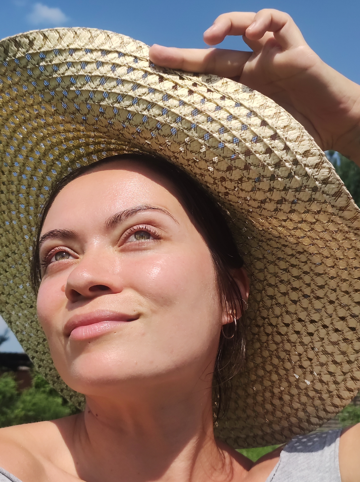 A close-up, eye-level, outdoor shot shows a young woman wearing a wide-brimmed straw hat against a bright blue sky. Her hand is visible on the hat, holding it in place. The woman is looking upwards and to the left, with a gentle smile on her face. Her skin is illuminated by the sun, highlighting her eyes and lips. Green foliage and the suggestion of a building are visible in the background, indicating an outdoor setting, likely in the city of Fokino, Russia.