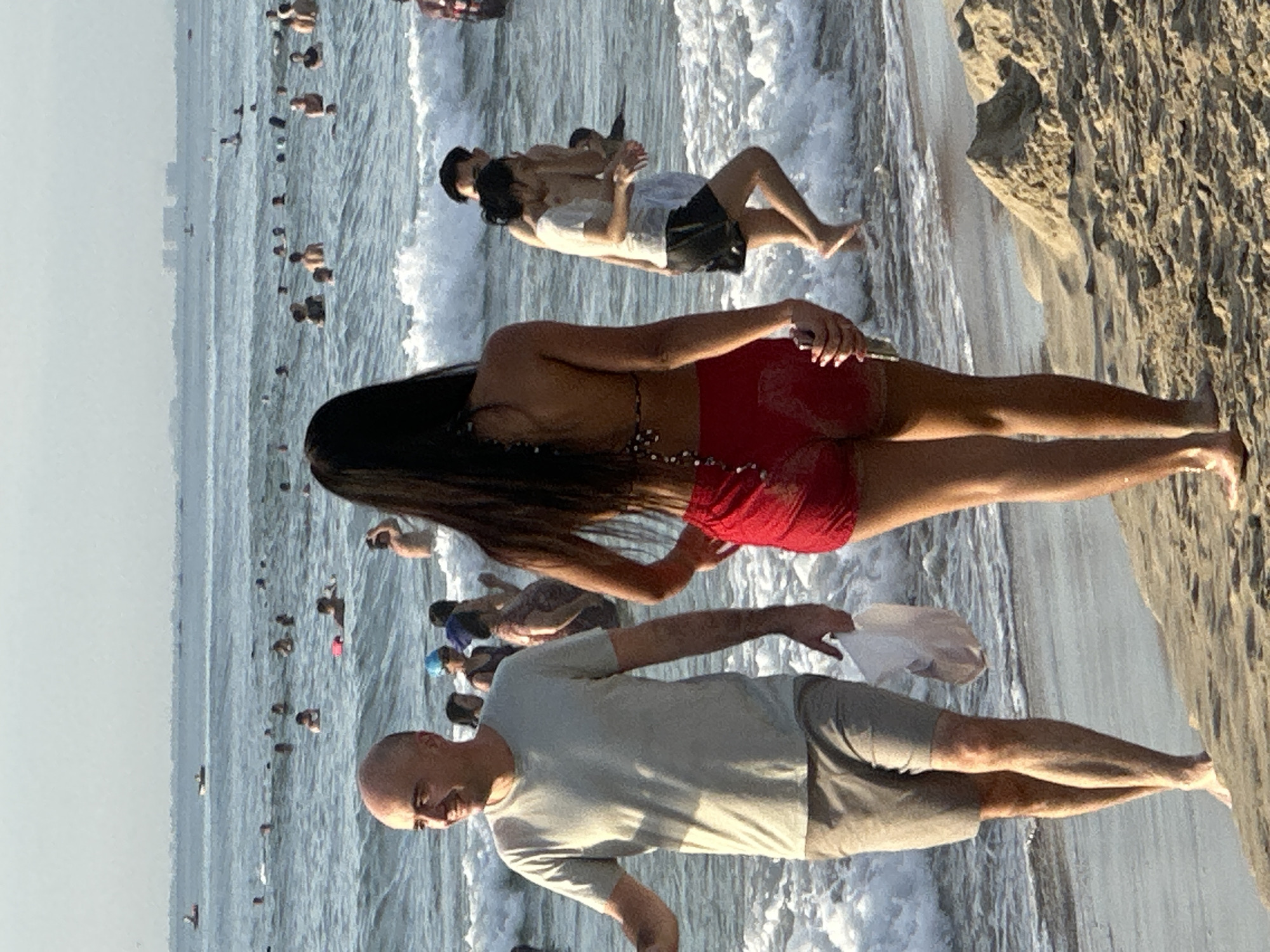 The image depicts a bustling beach scene in Da Nang, Vietnam, likely My Khe Beach, known for its expansive shoreline and clear waters. In the foreground, a bald man with light skin, wearing a light grey t-shirt and grey shorts, is walking on the wet sand, carrying a white plastic bag in his left hand. To his right and slightly ahead is a person with long dark hair, wearing a red bikini bottom and a patterned bikini top. Their back is to the camera, obscuring their face. The pair appear to be walking together along the waterline.
In the middle ground, the ocean is filled with numerous people swimming and wading in the water. The water is relatively calm with small waves breaking near the shore. Several individuals are visible from the waist up, enjoying the sea. Further in the background, a hazy skyline suggests the presence of buildings and possibly other coastal developments. The sky is a pale, hazy blue, indicating fair weather. The overall atmosphere is one of leisure and recreation, with many people enjoying a day at the beach.