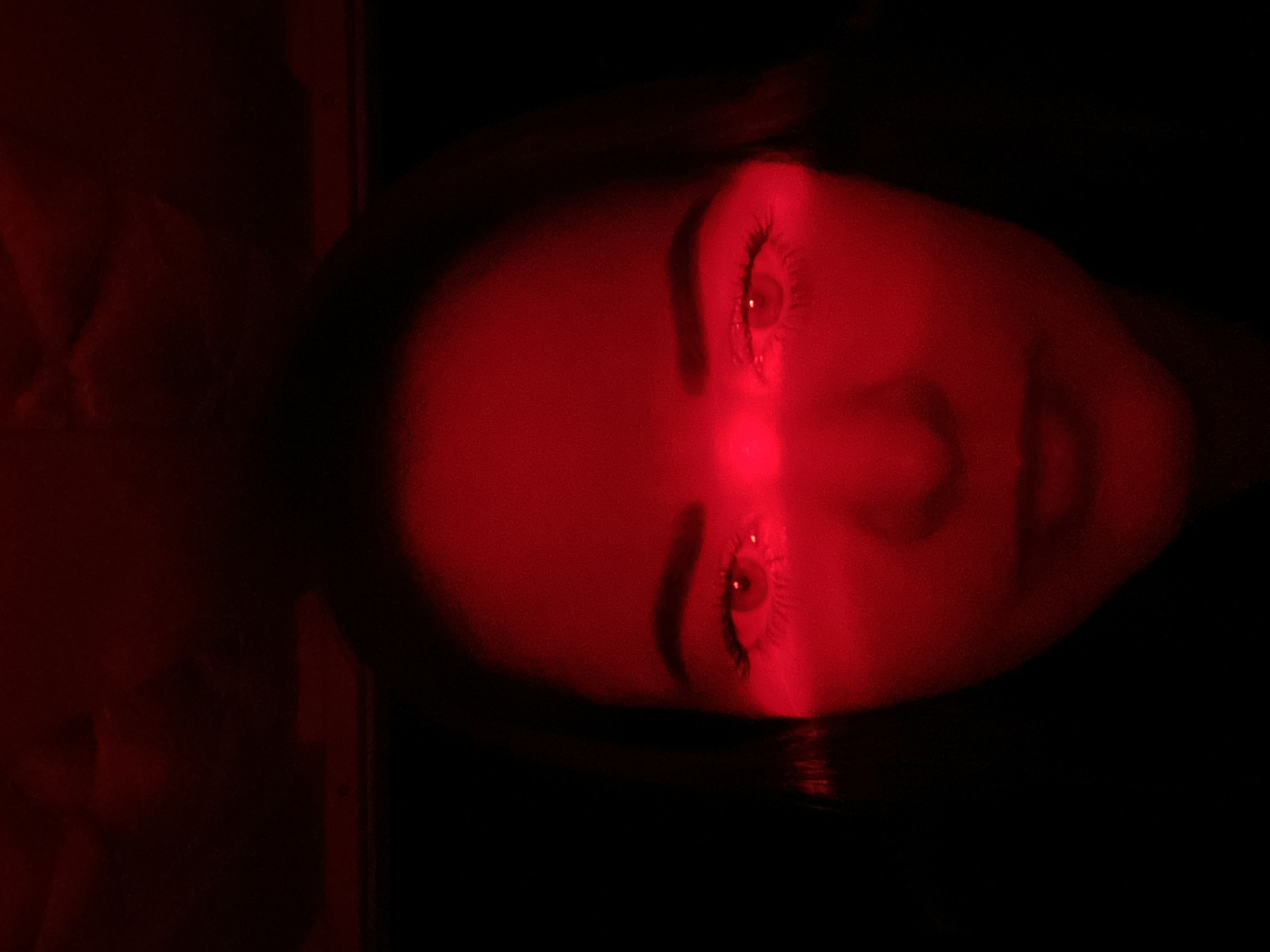 This media file presents a close-up, overhead view of a person's face, dramatically illuminated by a horizontal beam of bright red light. The subject, likely female due to visible eyelashes and facial structure, appears to be lying down, with their head positioned as if upright towards the top of the frame.
A distinct, narrow band of intense red light cuts across the face, illuminating both eyes, the bridge of the nose, and the upper cheeks. The eyes are open, with their pupils and irises reflecting the red light, making their natural color difficult to discern. Long, dark eyelashes are clearly visible. The skin, where not directly struck by the red beam, is shrouded in deep shadow, contributing to a high-contrast, almost silhouette effect against the vibrant red.
The overall environment is profoundly dark, suggesting an indoor setting with minimal or no ambient lighting other than the focused red source. The background is indistinguishable, fading into blackness around the edges of the face. The strong red light and deep shadows create a mysterious and intense atmosphere, highlighting specific facial features. No specific activity beyond being illuminated is evident. There are no discernible objects, location cues within the image, or visible text. The image is set in Yekaterinburg, Russia.