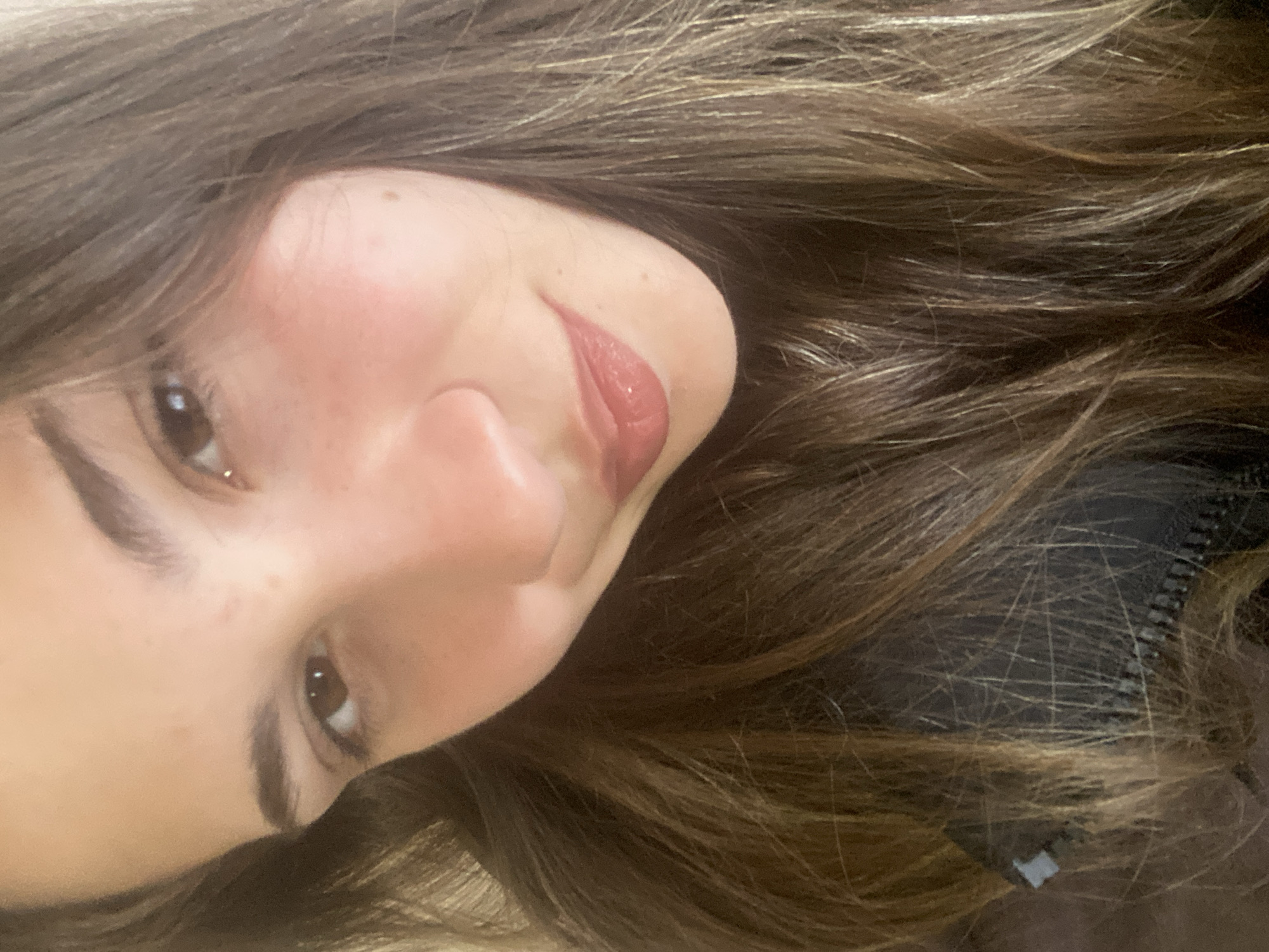 A selfie of a female with long, light brown hair and brown eyes. She has a light complexion, a medium-sized nose, and glossy lips. The background is out of focus, but a dark-colored garment with a zipper detail can be seen near the bottom right. The location is not discernible.