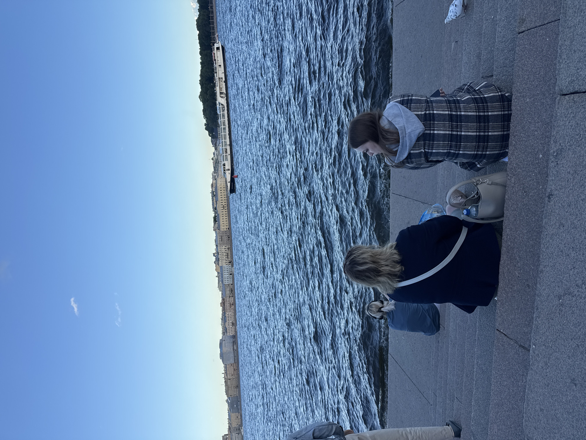 The image depicts a scenic view of the Neva River in Saint Petersburg, Russia, with the city's historic architecture visible in the background. Three individuals are seated on granite steps leading down to the water. The person closest to the foreground, viewed from behind, is a woman with blonde hair, wearing a dark blue jacket and a white strap across her shoulder. Next to her, also seated on the steps, is a young woman wearing a plaid jacket. Further back, another individual with blonde hair pulled into a ponytail is seated, facing the water.
The Neva River is a prominent feature, with choppy, dark blue water reflecting the clear, pale blue sky. A long, white passenger boat with several decks is navigating the river, heading towards the right of the frame. The cityscape across the river is lined with multi-story buildings displaying a variety of architectural styles. Trees and greenery can be seen behind the buildings on the far bank. A pigeon is visible on the steps in the lower right corner of the image. The overall atmosphere suggests a leisurely daytime scene, likely during the warmer months in Saint Petersburg.