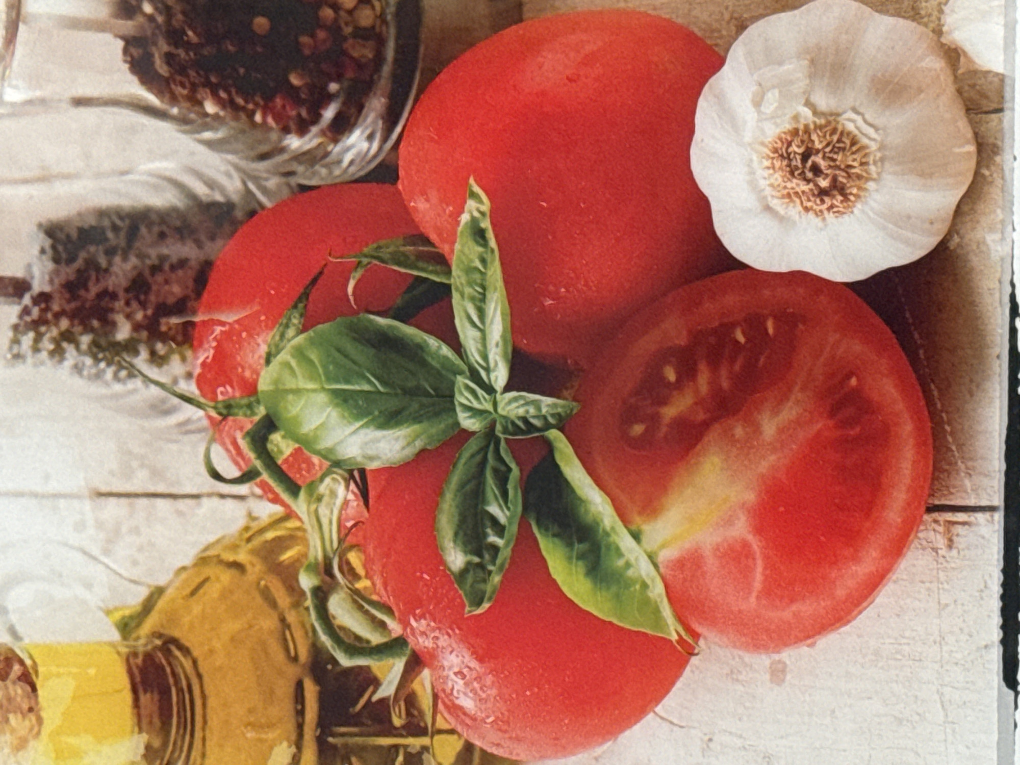 A close-up, eye-level, still life photograph features a collection of fresh ingredients arranged on a weathered wooden surface. In the foreground, a halved tomato reveals its juicy interior, next to a whole bulb of garlic. Behind the tomatoes, a glass bottle of golden olive oil stands prominently. Two clear glass spice shakers filled with peppercorns and crushed red pepper flakes are visible in the background, adding color and texture to the composition. The lighting is bright and natural, highlighting the vibrant colors and textures of the food items.