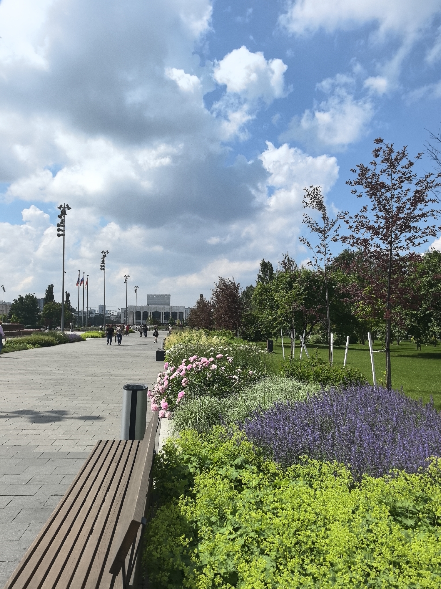 The image depicts a park area in the city of Perm, Russia, with a paved walkway in the foreground, lined with benches and planters. Several people are walking along the pathway. In the background, a large building, possibly a cultural center or museum, is visible. The sky is partly cloudy, with scattered white clouds against a blue backdrop. The park features manicured lawns, trees, and flowerbeds, creating a pleasant urban green space.