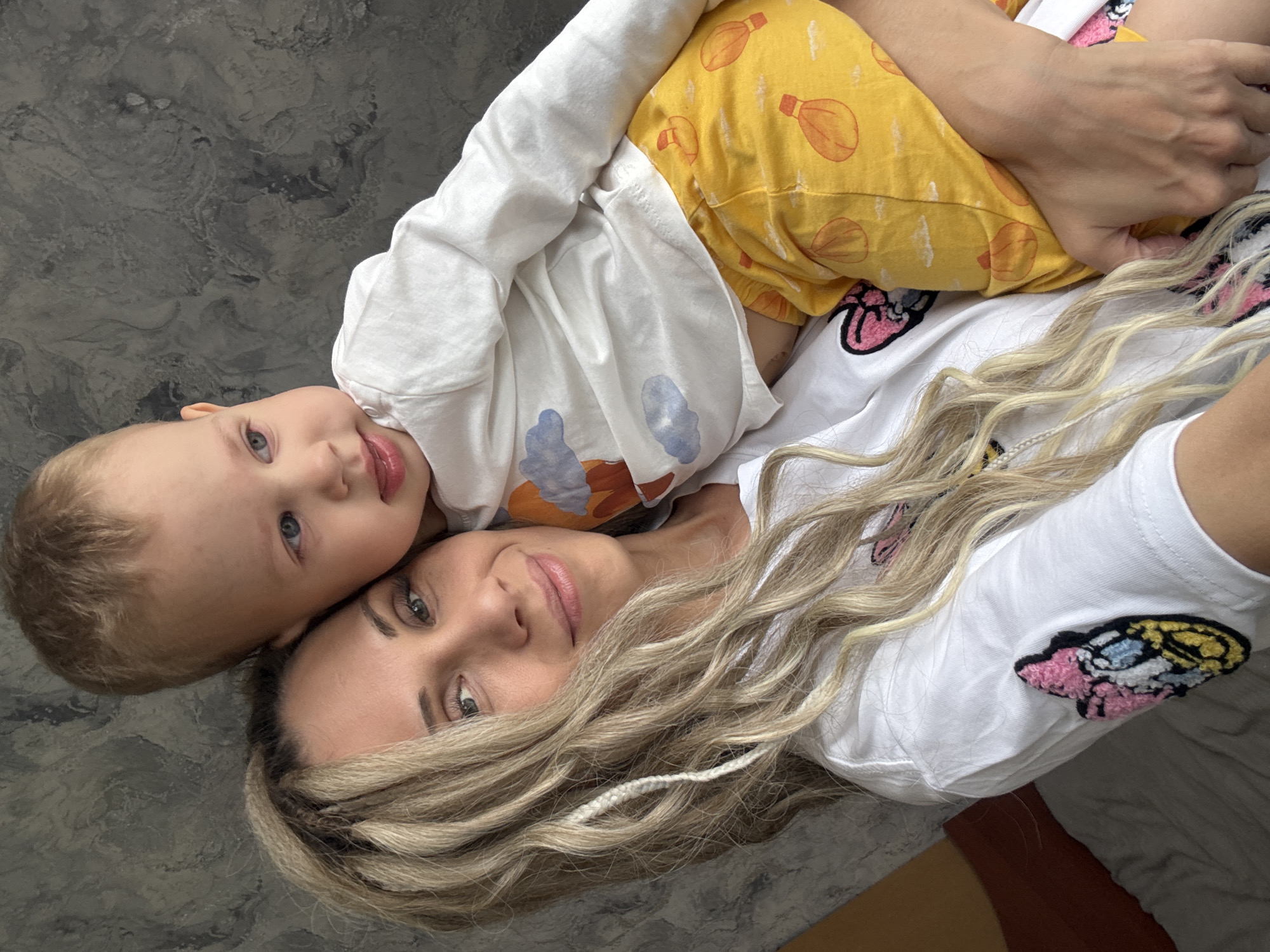 A woman and a young child are lying down, close together, in what appears to be an indoor setting. The woman is lying on her back, with her head tilted towards the camera, showcasing her face with light makeup and wavy blonde hair. A single white braid is visible in her hair. She is wearing a white t-shirt with a colorful embroidered patch. The child is positioned on top of the woman, with their head resting near her head, and their body across her chest. The child has fair skin, blonde hair, and blue eyes. They are wearing a white long-sleeved shirt over a yellow shirt printed with hot air balloons. The background has a gray, textured surface, and a sliver of brown flooring is visible in the bottom corner of the frame. The location is in the city of Unknown, Unknown.