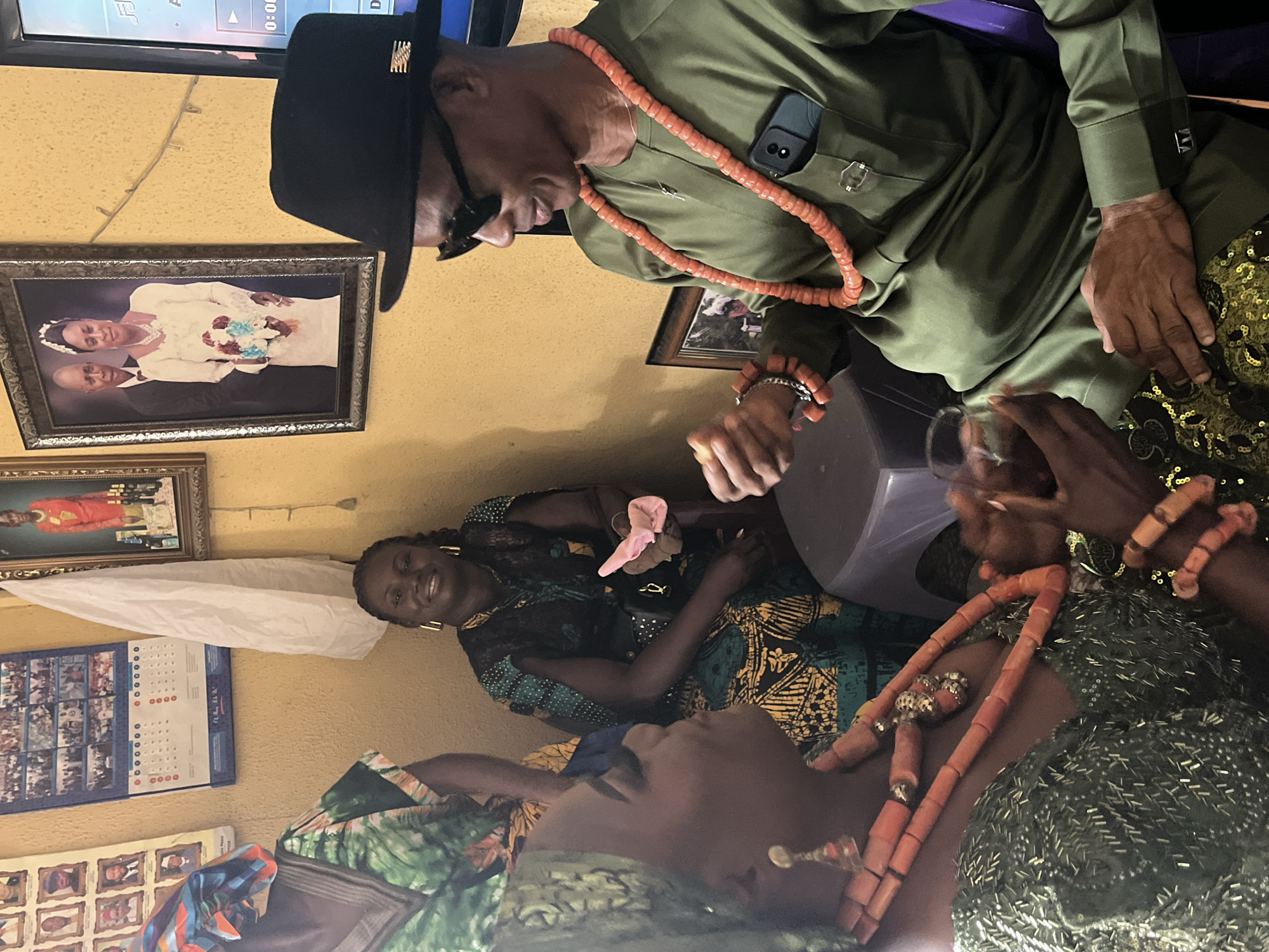 In an interior setting, possibly a residence in Warri, Nigeria, a man in an olive green traditional attire, black hat, and sunglasses, with coral beads around his neck and wrist, stands beside a woman in a patterned dress and another woman in a sequined dress with coral beads. The man is looking down, possibly interacting with the women. The woman in the patterned dress is smiling. On the wall are framed photos, including one of a couple in wedding attire, and a calendar. A television is mounted high on the wall. A white cloth hangs from the wall.