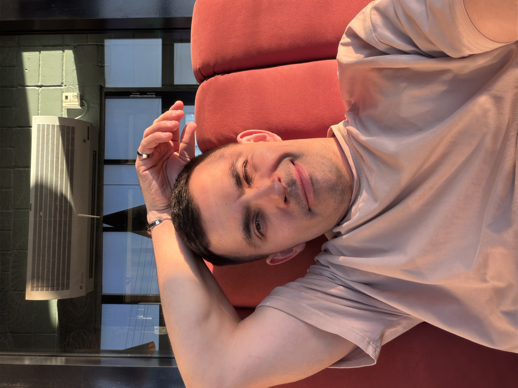 A fair-skinned man is reclining on an orange upholstered surface. He is wearing a light brown t-shirt, a ring on his left ring finger, and a bracelet on his left wrist. His right eye is partially closed. Behind him, there is a window with a view of an air conditioning unit and a brick wall in the city of Izhevsk, Russia.