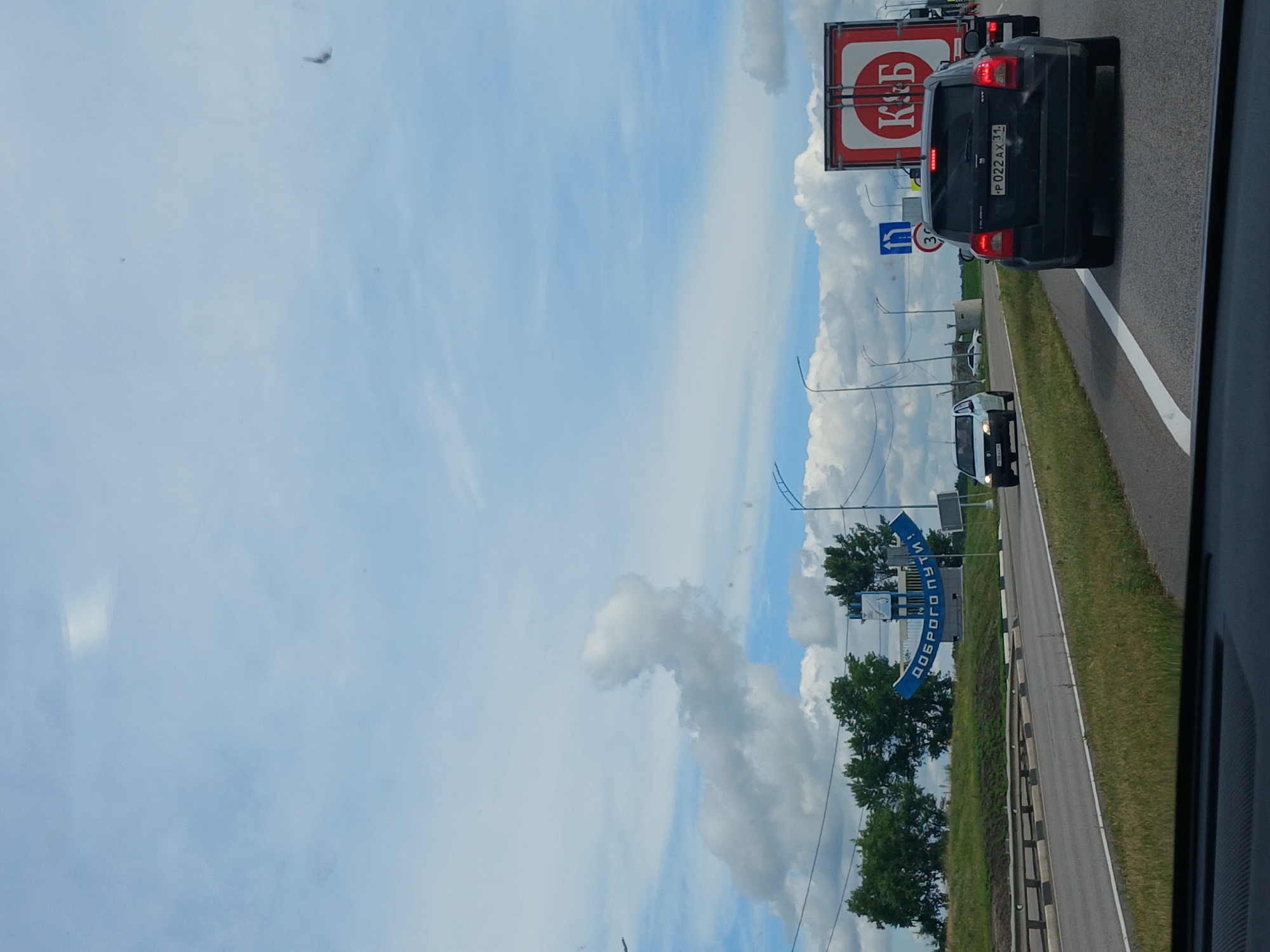 This is a horizontal, eye-level, outdoor, daytime, shot of a highway in Russia. The image is taken from the interior of a vehicle, looking out through the windshield. In the foreground, the dashboard of the vehicle is visible, a dark grey color with a textured surface.
The highway itself is paved and has clear white lane markings. There are two lanes of traffic visible, with grass and vegetation lining the sides. A metal guardrail separates the lanes from the shoulder.
In the middle ground, there are several vehicles on the highway. A black SUV is in the right lane, with its brake lights illuminated. A large red semi-trailer truck with a white circular logo containing the letters "К&Б" is to the right of the SUV. In the left lane, a white car is visible further ahead. Further to the left, a large blue archway sign is visible on a grassy embankment, with text in Russian that reads "ДОБРОГО ПУТИ!". Behind the sign, trees and some structures are visible.
In the background, the sky is a light blue with scattered white, puffy clouds. The overall scene depicts a typical highway view with traffic and roadside elements.