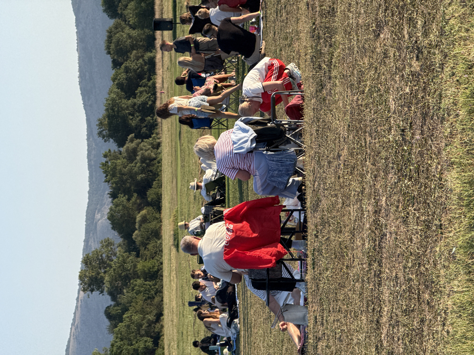 In Orlinivka, Ukraine, a group of people are gathered outdoors on a grassy field, likely for an event. The scene is bathed in the warm, late afternoon sun. Many individuals are seated in foldable chairs, while others are on blankets on the ground, suggesting a relaxed, informal gathering. In the foreground, a man is seated in a chair, a red jacket with a Coca-Cola logo draped over it. To his right, a young person in red athletic wear is bent over, appearing to be retrieving something from a bag. Further back, a woman in a striped shirt is seated, looking towards the center of the gathering. In the middle ground, a woman in a white top and denim shorts walks past other seated individuals, carrying a pair of flip-flops. More people are visible in the background, some standing, others seated, all facing towards a common point of interest that is not fully visible in the frame. A black speaker is placed on a stand in the far right background, hinting at a possible performance or presentation. The environment is characterized by a wide, open grassy area bordered by a line of trees and rolling hills in the distance under a clear sky.