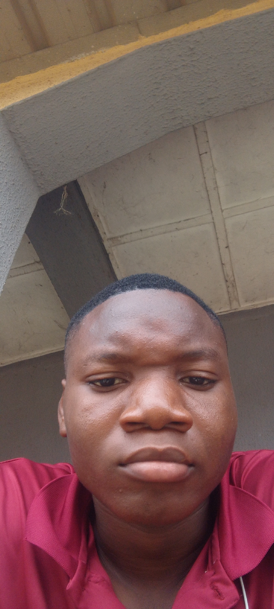 This is a close-up, eye-level, selfie-style photograph of a young Black person. The subject is positioned centrally in the frame, with their face taking up most of the space. They have dark, short, neatly cut hair, dark eyes, and prominent cheekbones. Their skin appears smooth and has a warm, brown tone. They are wearing a collared, maroon shirt. The background of the image is composed of structural elements, possibly a covered outdoor space. A concrete beam or overhang is visible at the top of the frame, with a lighter colored, possibly tiled ceiling extending beyond it. A small piece of dried vegetation is hanging from the overhang. The lighting suggests it is daytime, but the exact time is unclear due to the overcast or shaded conditions. The subject's expression is neutral, and they appear to be looking directly at the camera. The setting seems to be an outdoor or semi-outdoor location, perhaps a porch or walkway, with architectural features providing a backdrop. The location context is Suleja, Nigeria. There is no visible text in the image.