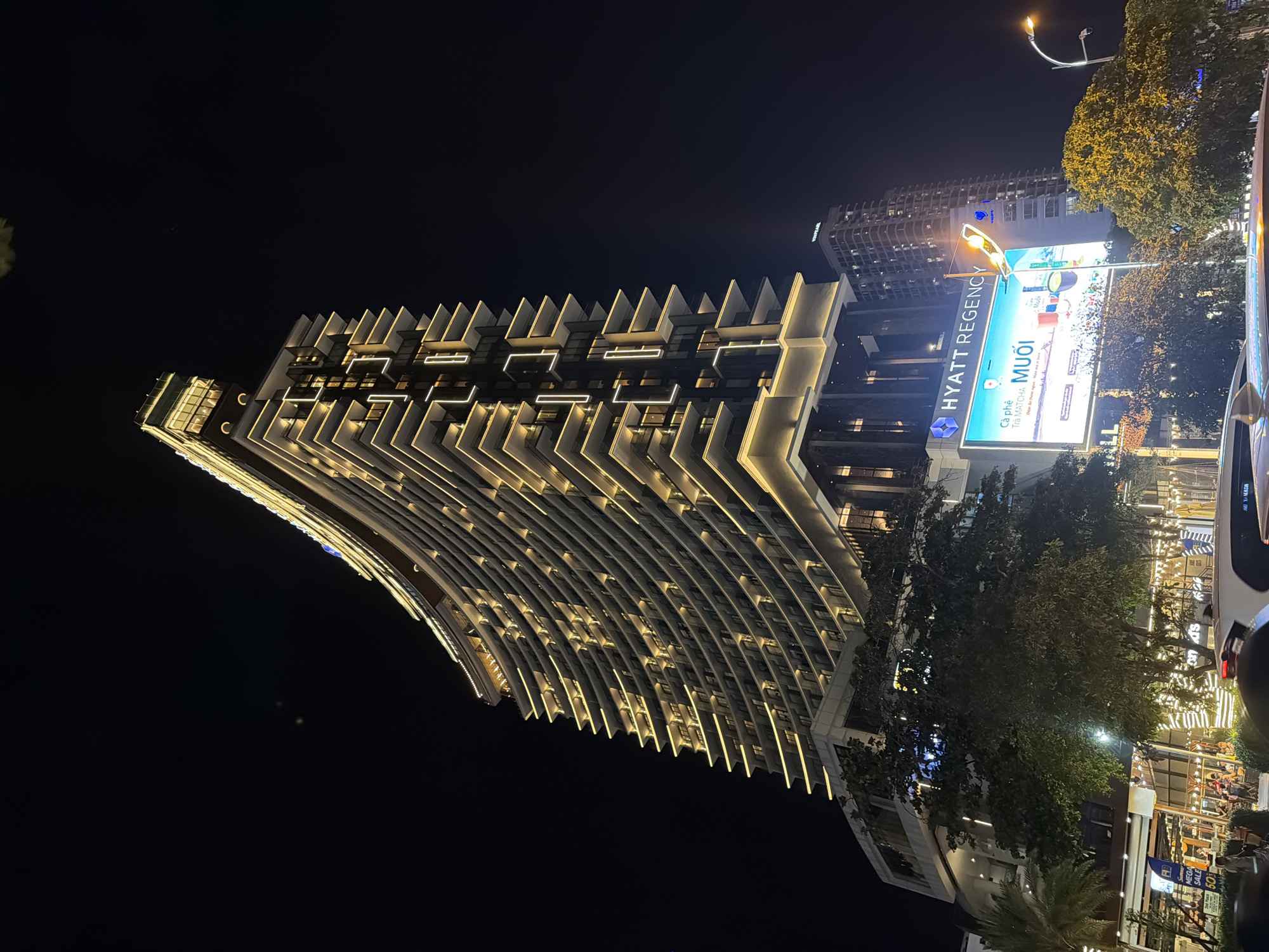 The image shows the Hyatt Regency Nha Trang hotel, a modern high-rise building illuminated at night. The hotel's curved facade is lined with balconies and features warm yellow lighting along its edges and on individual balconies. A large digital billboard below the hotel displays an advertisement in Vietnamese, featuring a matcha drink. To the right of the Hyatt Regency, another tall building is visible. In the foreground, there are trees, streetlights, and the tops of vehicles, suggesting a street-level view in an urban environment. The overall scene is vibrant and illuminated, typical of a city at night.
