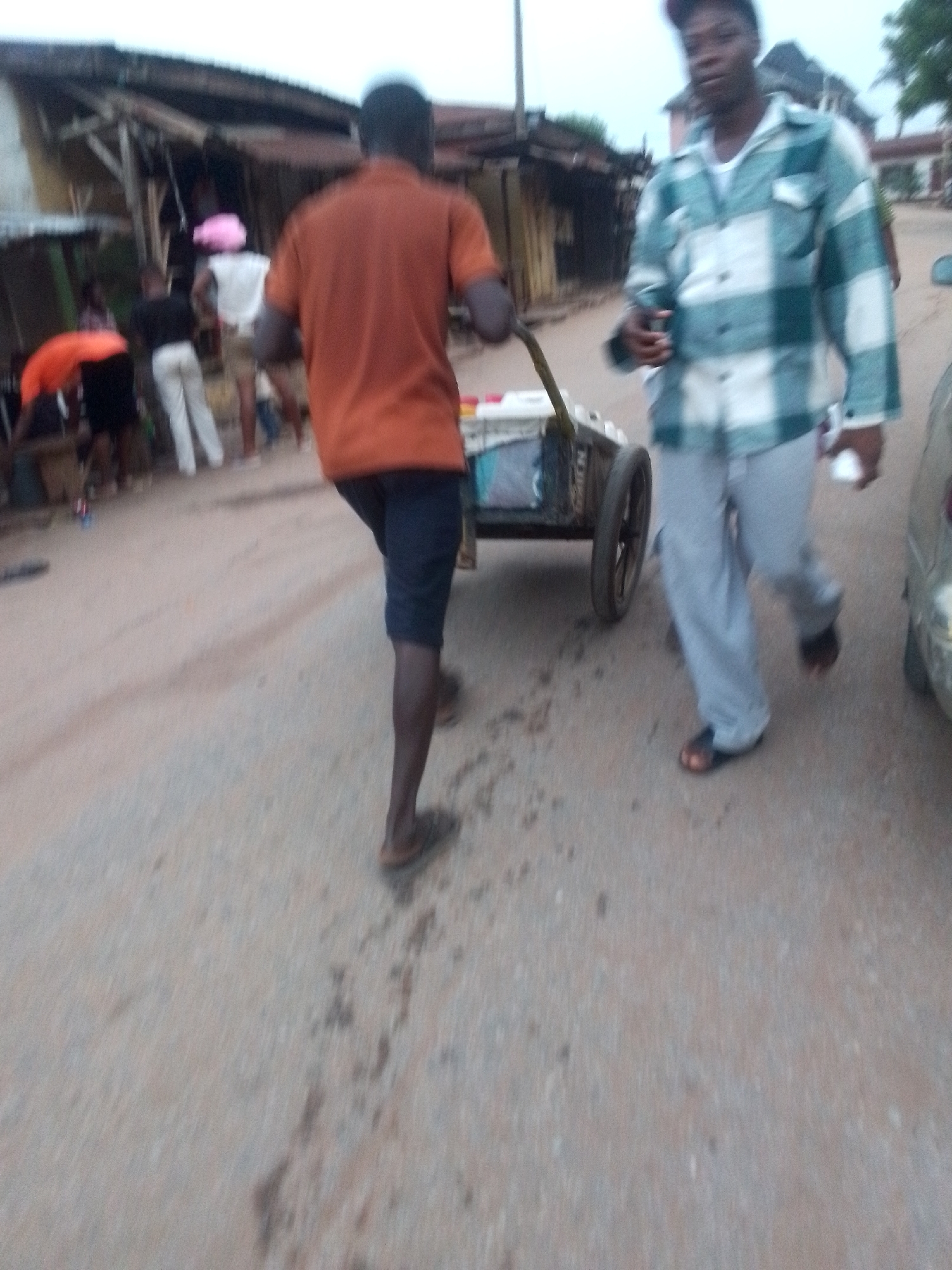 The image shows a street scene in Onitsha, Nigeria. In the foreground, a man in a teal and white plaid shirt and grey pants walks towards the viewer, holding an object in his hand. To his left, another man in an orange shirt and dark shorts pulls a cart. In the background, several people stand near what appears to be a row of shops or stalls with corrugated metal roofs.
