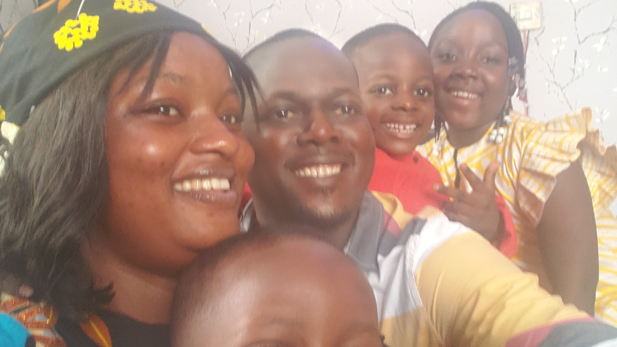 The image depicts a family portrait featuring five individuals, possibly taken indoors given the muted, patterned wallpaper in the background. The scene is set in Barden Yana, Nigeria.
In the foreground, a woman on the left is close to the camera, smiling with her mouth open. She is wearing a dark headscarf with yellow and orange floral patterns. Beside her, a man is also smiling, his face filling the center of the frame. He is wearing a collared shirt with horizontal stripes of grey, white, and yellow. Behind the man and slightly to his right, a young child in a red shirt is smiling broadly. To the far right, a young woman with braided hair, wearing a yellow and white checkered dress with ruffled sleeves, is also smiling and making a peace sign with her hand. Partially visible in the very front, the head of another child is present, obscuring the lower faces of the adults in front. The overall impression is a candid, happy moment captured by the family.