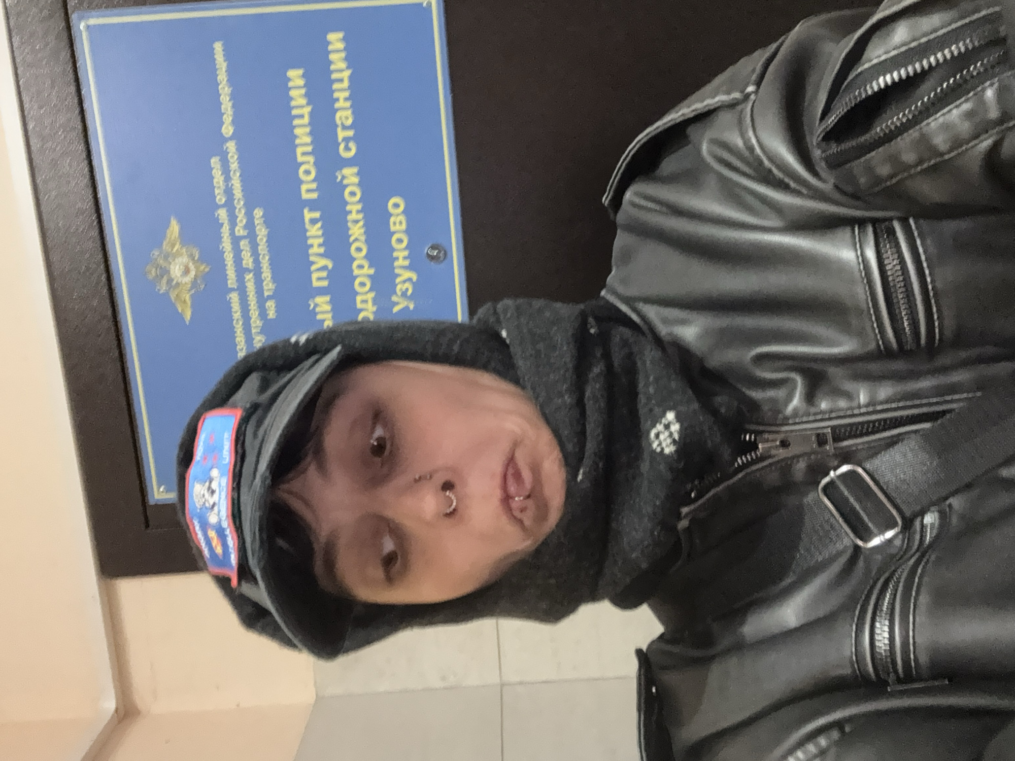 An individual is captured in a selfie-style photograph, shown from the chest up, with their head and upper body rotated. They are wearing a black leather jacket with multiple prominent zippers, a dark gray hoodie underneath, and a dark cap adorned with a colorful patch featuring a character resembling Mickey Mouse. The person has facial piercings, including a septum ring and a labret piercing, and is making a quirky expression with their tongue slightly extended and eyes looking upwards to the side.
In the background, a blue rectangular sign with text in Cyrillic script is clearly visible. An emblem, possibly a coat of arms related to law enforcement, is also present on the sign. The text, partially obscured but discernible through OCR and context, translates to "Ryazan Linear Department of Internal Affairs of the Russian Federation on Transport" and "Police Point of Uzunovo Railway Station". This identifies the setting as an indoor space, specifically a police point at a railway station in Uzunovo, Russia. The surroundings appear to be a simple wall, suggesting a public or official building.