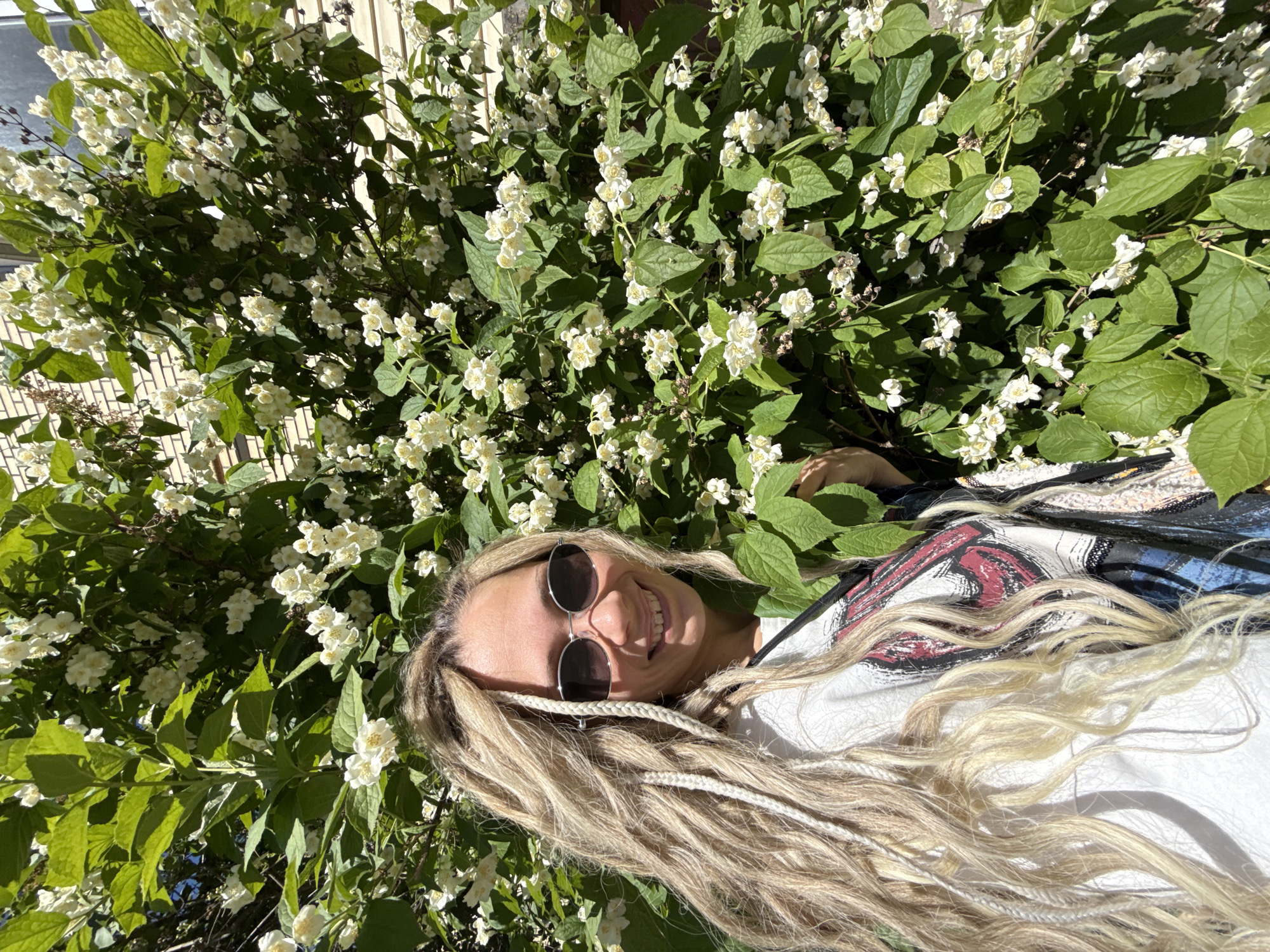 The image depicts a young adult female standing outdoors in Rusanovo, Russia, next to a large, blooming shrub. The individual is positioned in the left foreground, facing forward and smiling towards the camera. She has long, blonde hair styled with dreadlocks, some of which are braided with white strands. She wears oval sunglasses with silver frames and a white short-sleeved top featuring a multi-colored graphic design. A black strap, consistent with a bag or camera strap, is visible over her left shoulder. Her left hand is touching the leaves of the shrub. The shrub, which occupies the central and right portions of the frame, is dense with vibrant green leaves and numerous clusters of small, white, multi-petaled flowers with yellow centers. In the background, behind the foliage, sections of a building are discernible, showing light-colored brickwork and horizontal siding, along with a window. The scene is illuminated by direct sunlight, indicating a clear day.