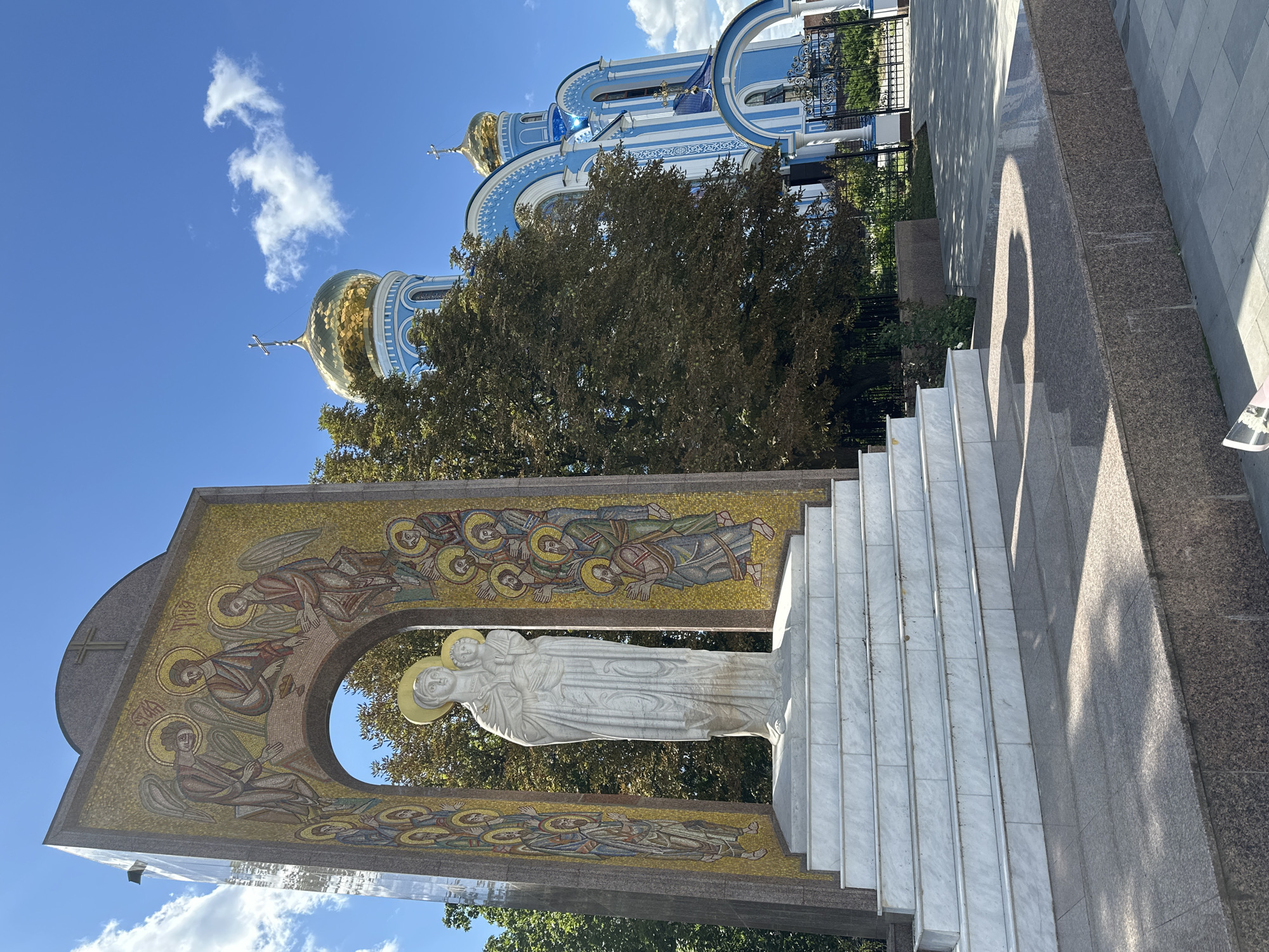 The image depicts an outdoor scene in the city of Zhovte, Ukraine, featuring a religious monument and a church. In the foreground, a white marble statue of the Virgin Mary holding the infant Jesus stands on a tiered white marble base. This statue is framed by an arched structure adorned with a gold mosaic depicting angels and religious figures, with a cross at the apex. The mosaic also features a side panel with a reflective, possibly glass or polished stone, surface. Behind the monument, a large deciduous tree with dark green foliage is visible. To the right of the tree and monument, a blue and white Orthodox church with golden domes is prominent. The church has intricate architectural details, including arched windows and decorative white trim against the blue walls. A decorative black metal fence borders the church property. The ground in front of the church appears to be paved with light gray tiles and darker granite. The sky above is bright blue with scattered white clouds. The lighting suggests it is daytime, likely with the sun casting shadows on the ground. There are no visible people in the image.