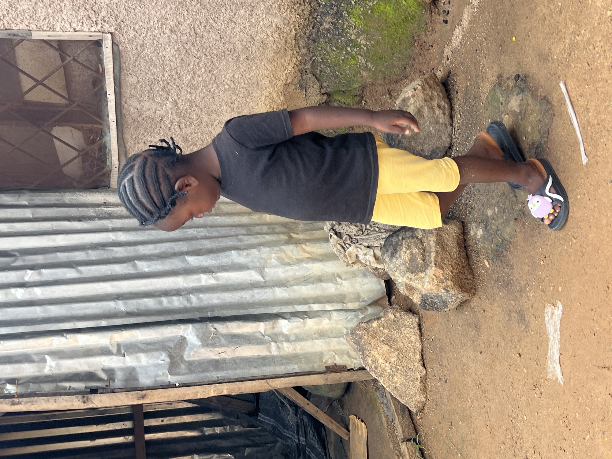 A young Black child, seen from the side, walks on a dirt path in Jos, Nigeria. The child wears a black t-shirt and bright yellow shorts, and a pair of black flip-flops with a pink cartoon owl design. The background features a corrugated metal structure on the left and a textured, tan-colored wall on the right. A window with a diamond pattern is visible in the wall. Several large rocks are scattered on the ground. The overall setting suggests an outdoor area, possibly a residential compound or a less developed urban environment.
